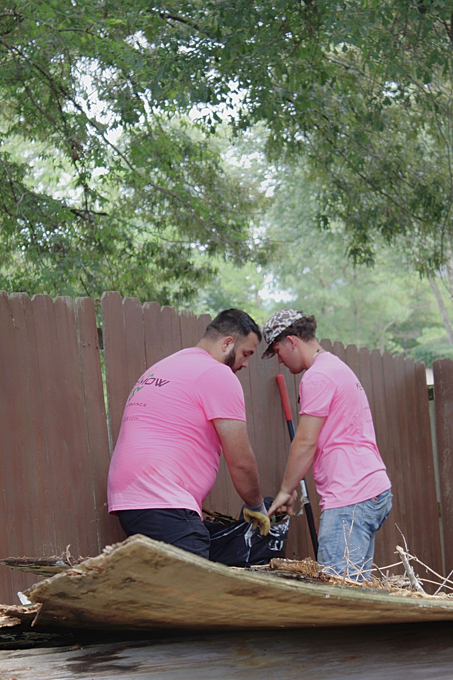 Two people in pink shirts work together to clear debris in a fenced backyard.