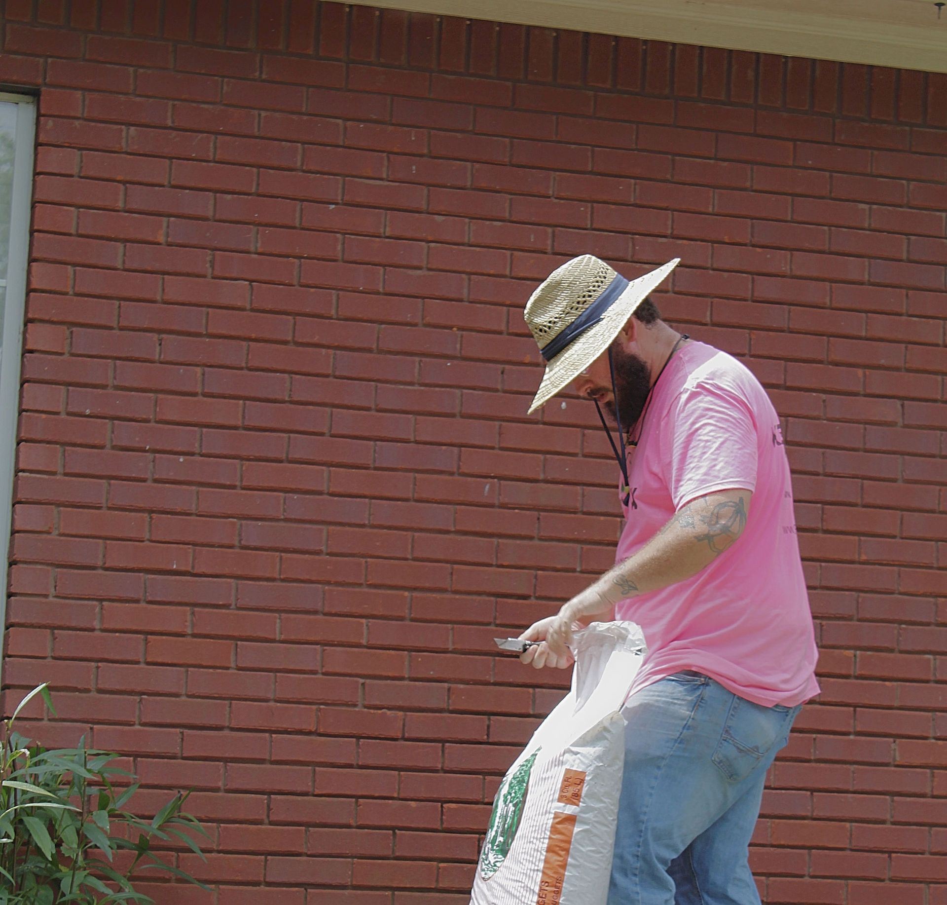Person in a pink shirt and straw hat working beside a red brick wall, holding a white bag.