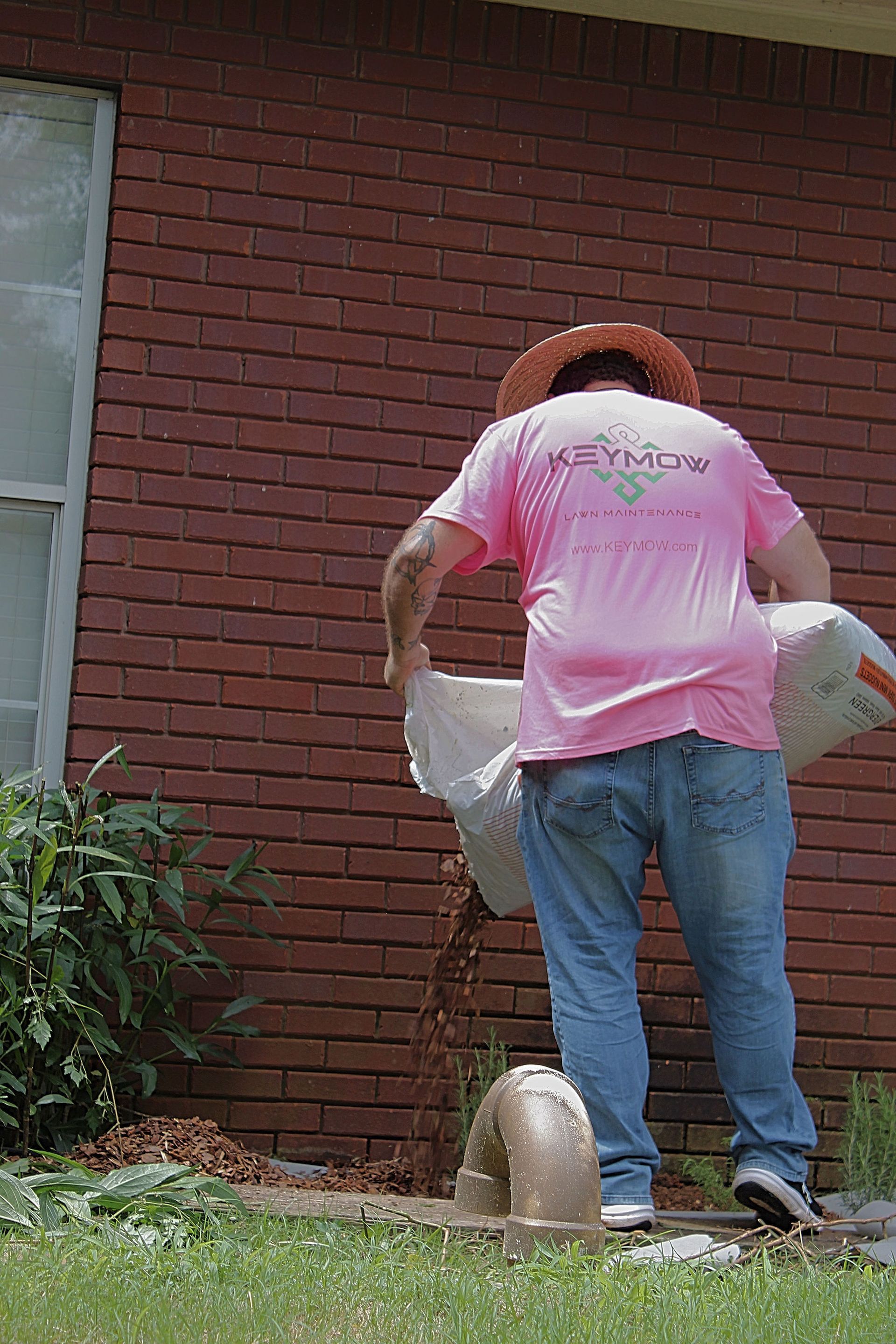 Person in a pink shirt carrying bags, standing by a brick wall with a garden hose and bucket.