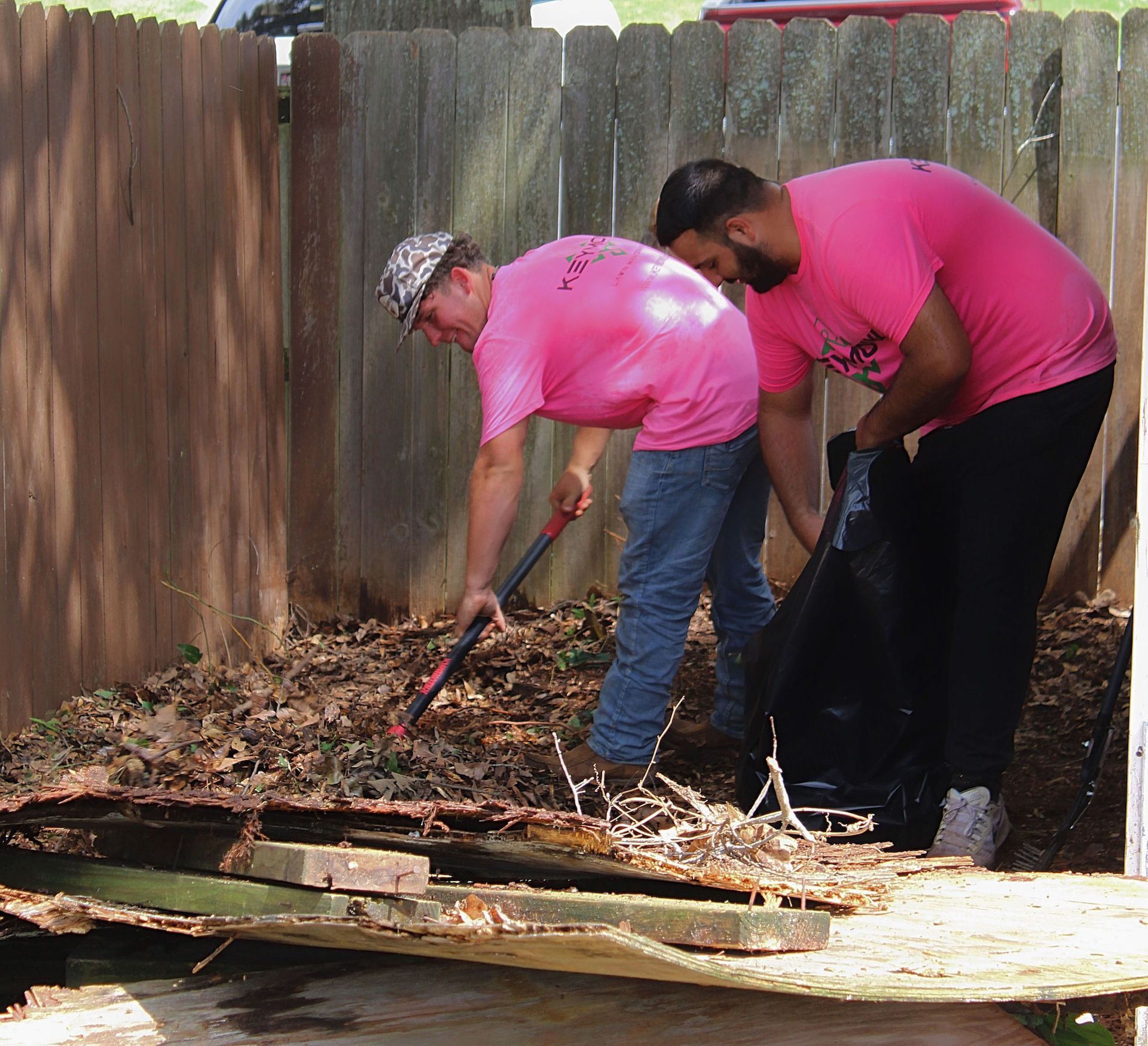 Two people in pink shirts clearing leaves and debris beside a wooden fence in a backyard.
