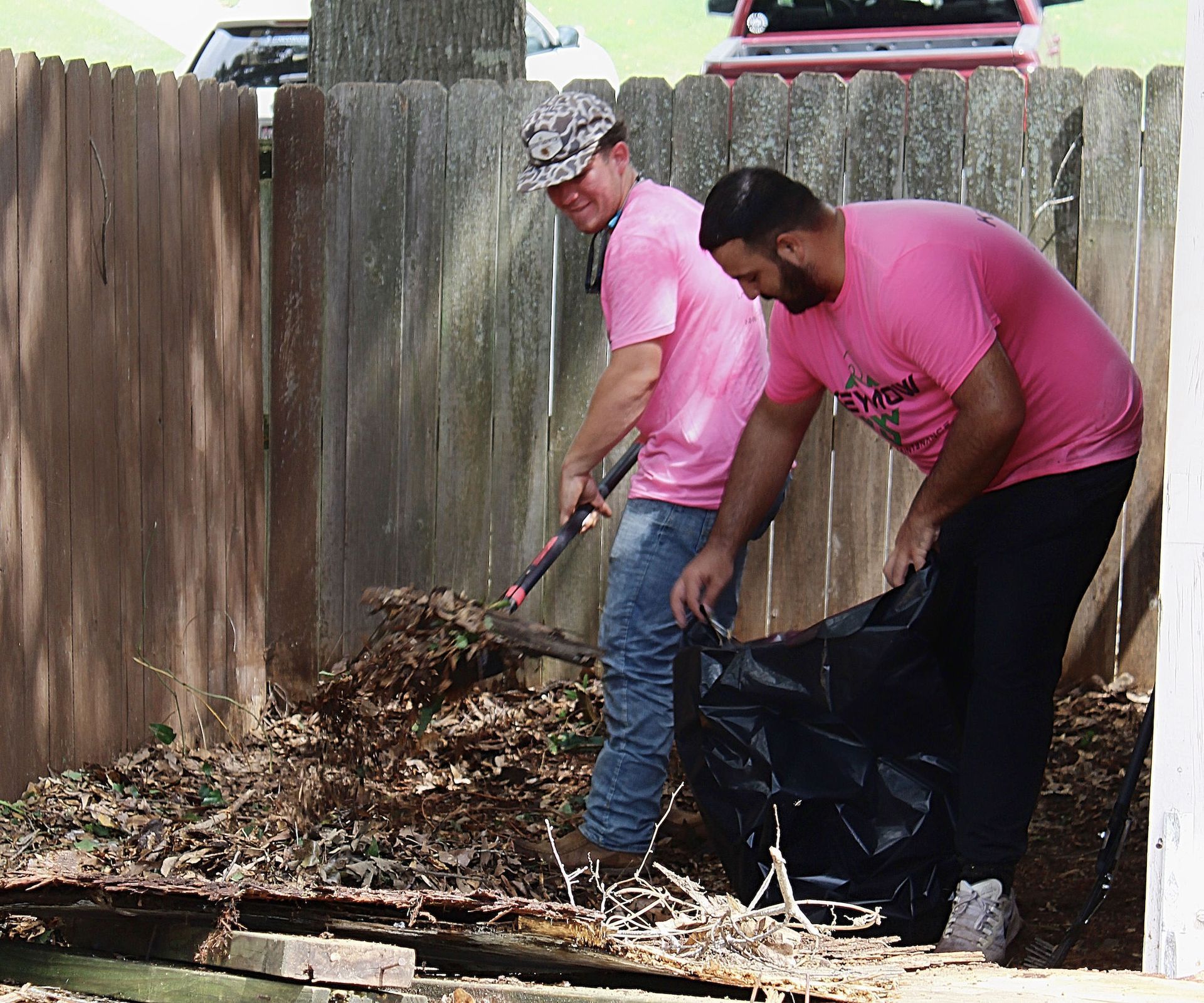 Two people in pink shirts raking leaves into a black bag in a fenced backyard.