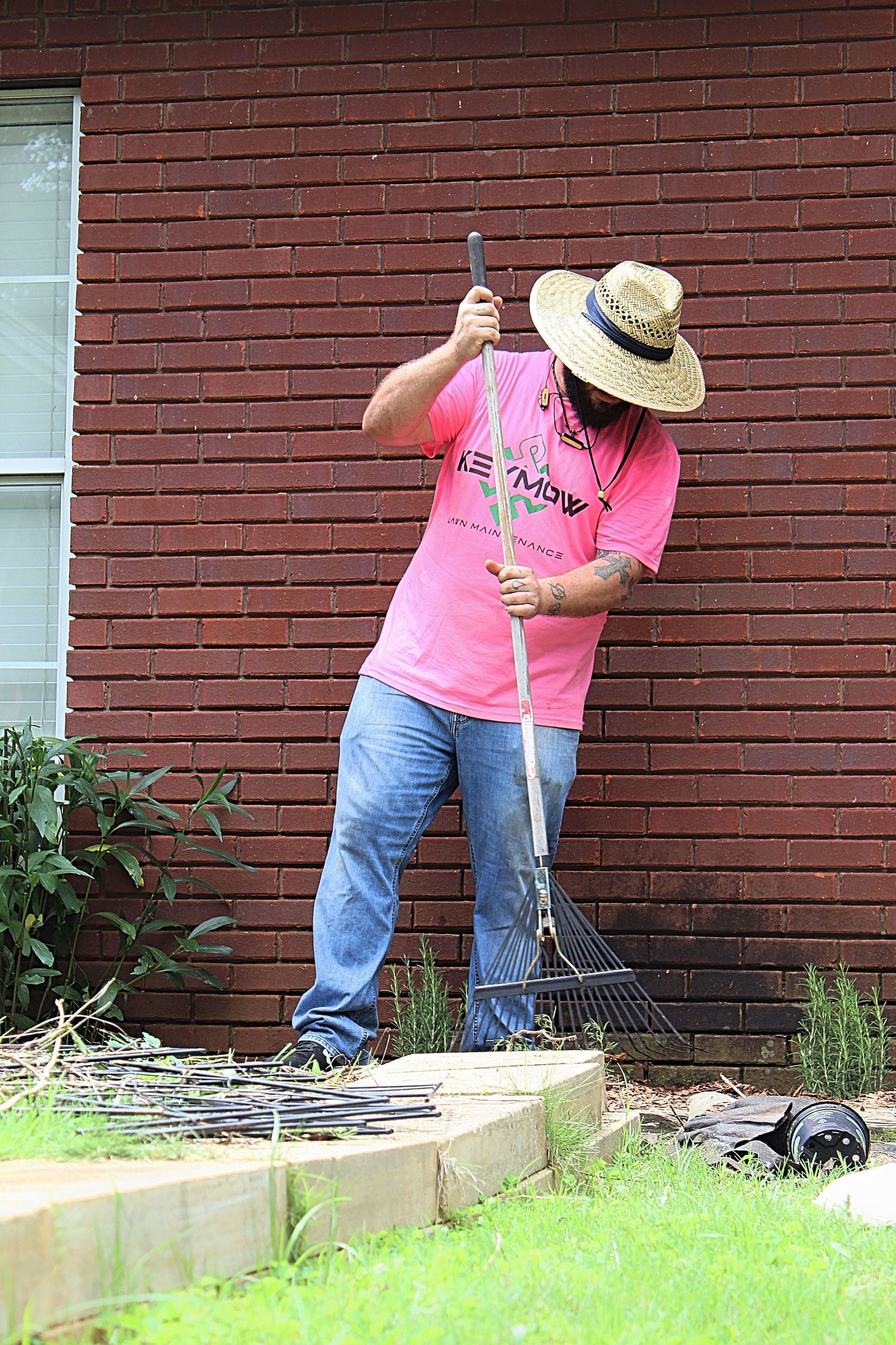 Person in pink shirt and straw hat using a shovel in a yard beside a brick wall.