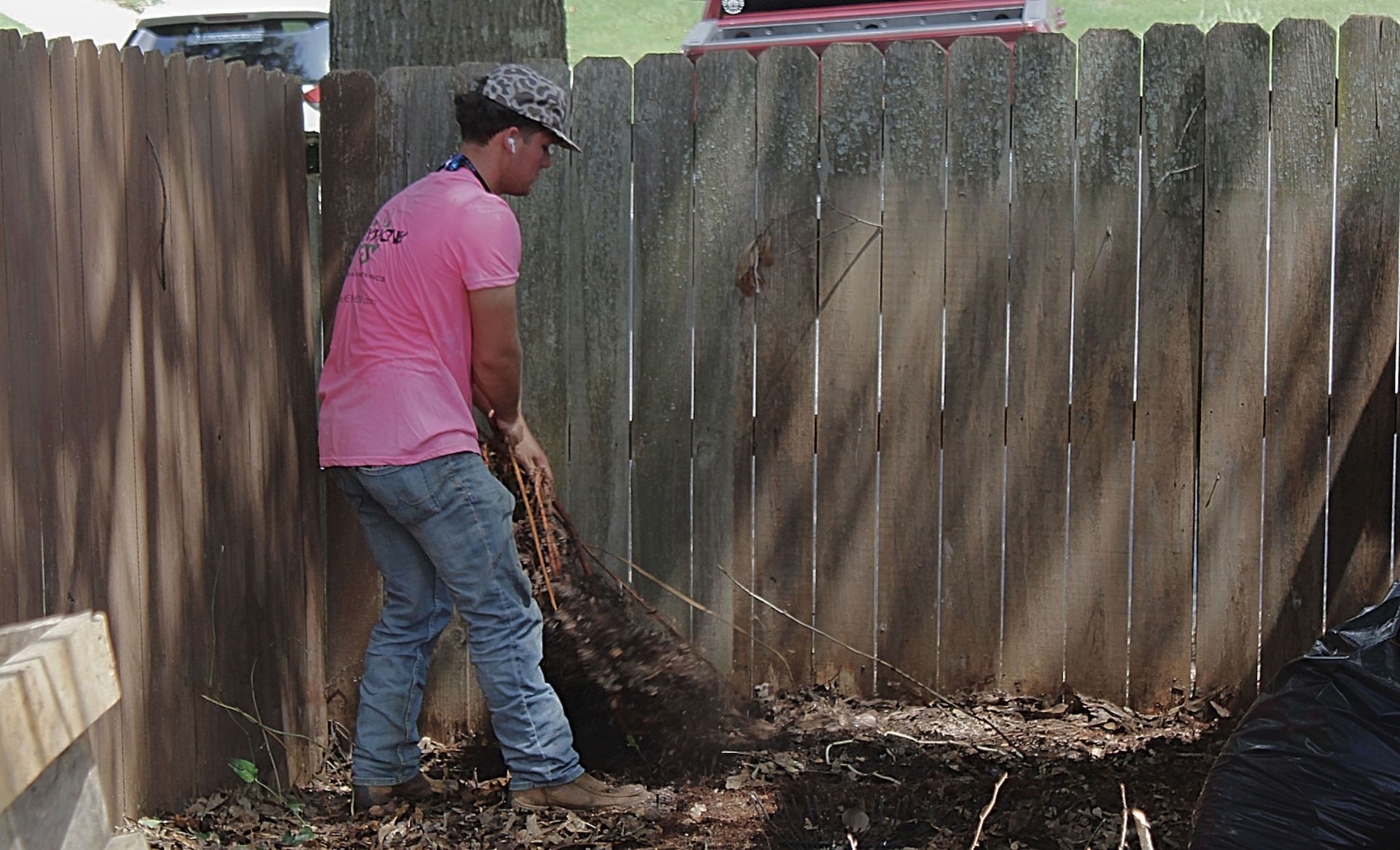 Person in a pink shirt and cap shoveling debris along a wooden fence in a yard.