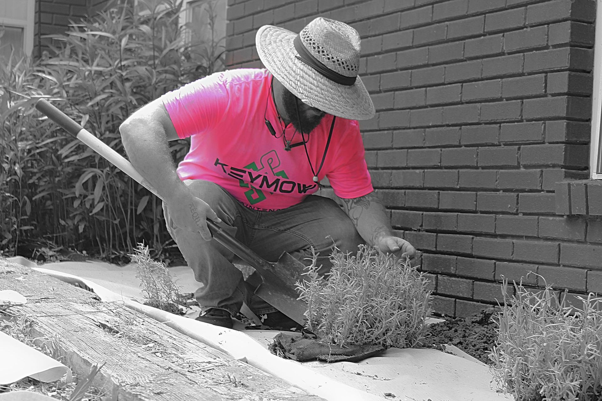 Person in a pink shirt and straw hat kneels weeding beside a brick wall and garden bed.