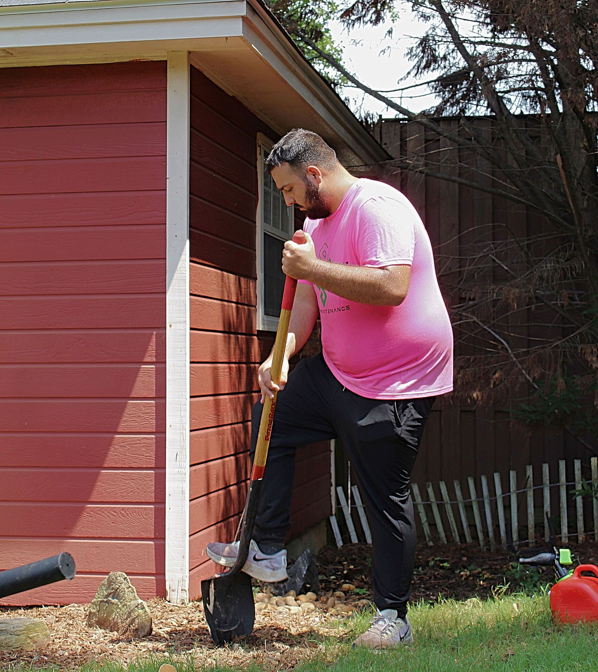 Person digging in a backyard beside a red shed, wearing a pink shirt and black pants.
