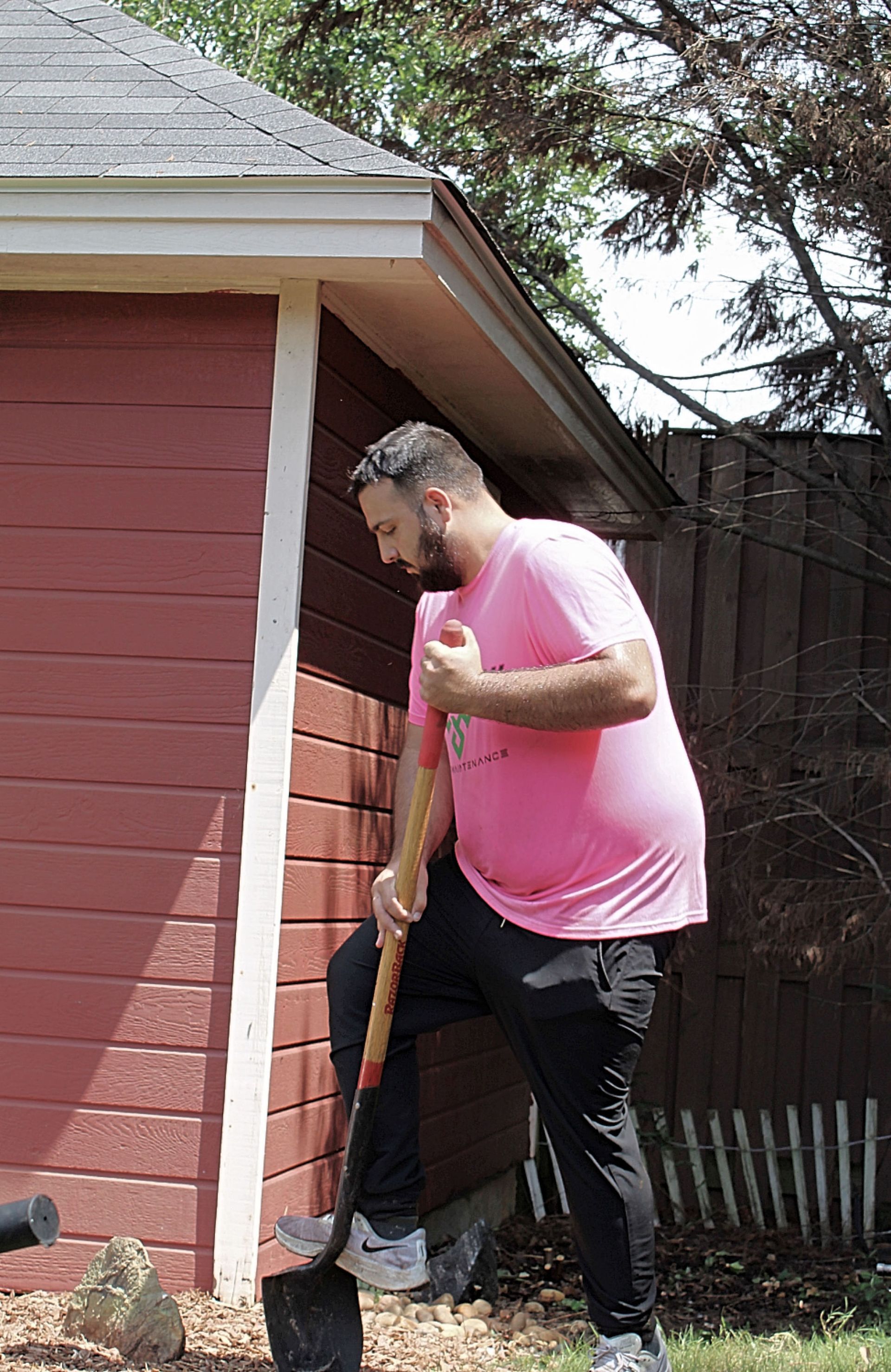 Person in a pink shirt using a shovel beside a red house and wooden fence.