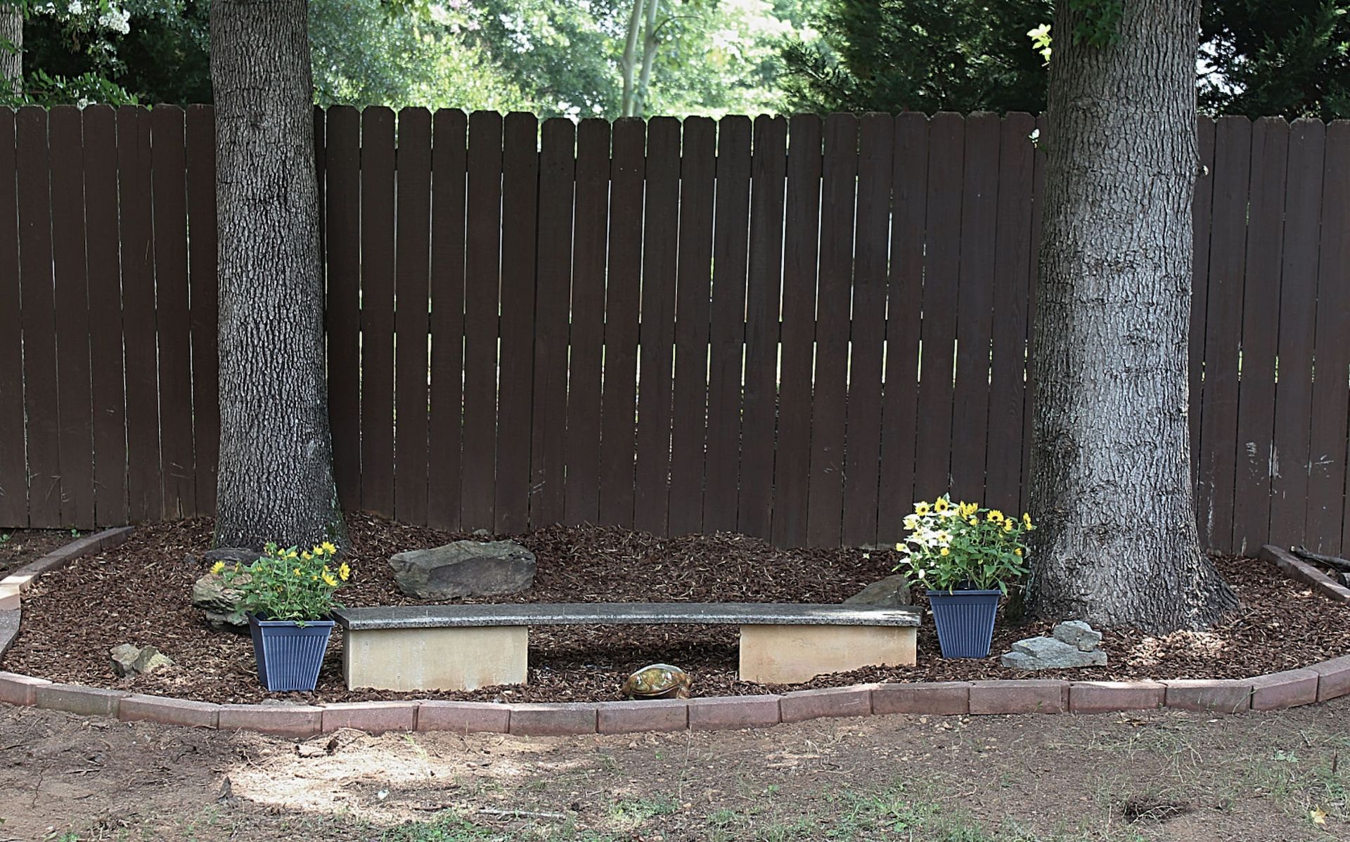 Small backyard garden bed with potted flowers, trees, and a wooden fence.