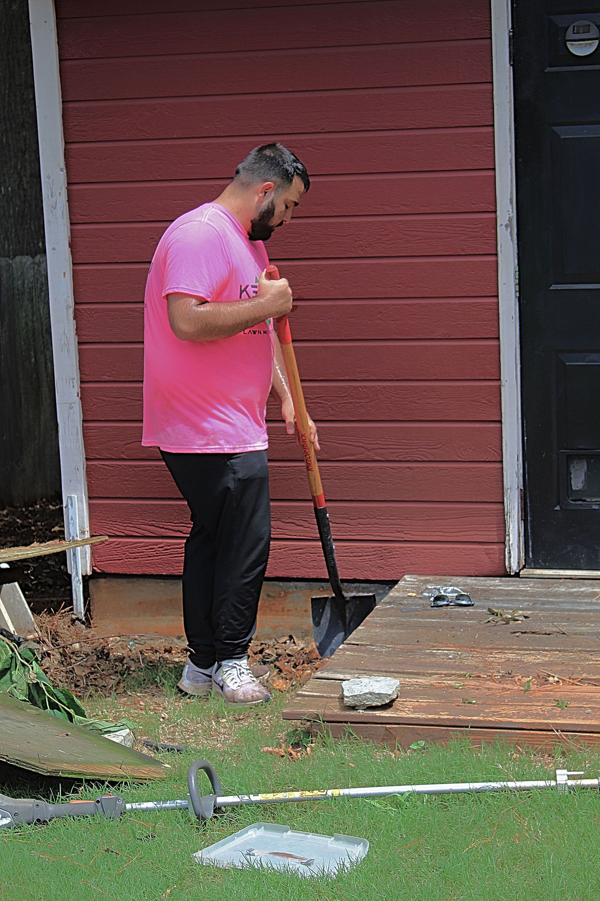 Person in a pink shirt using a shovel to clear debris on a porch outside a red house.