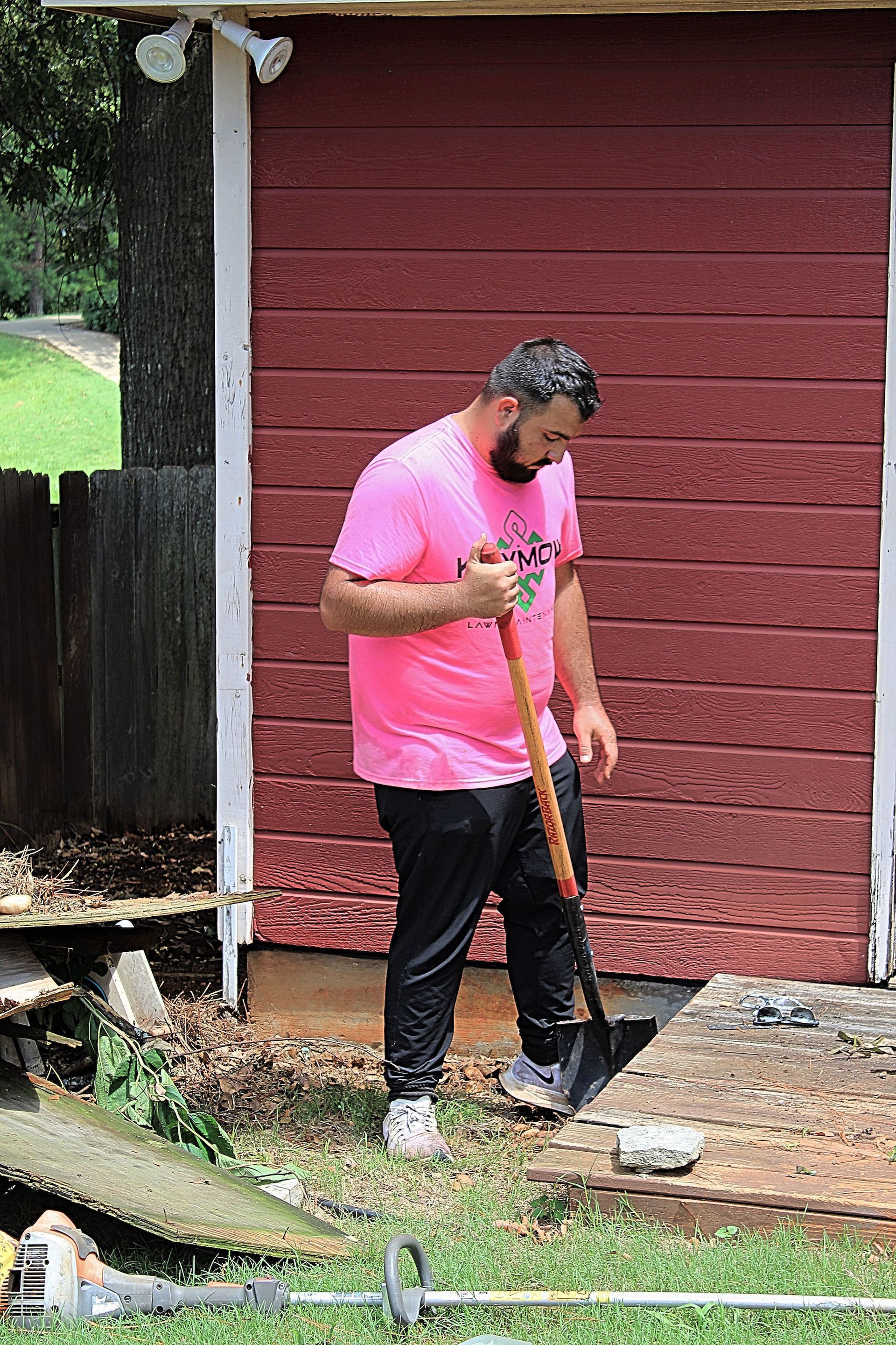 Person in a pink shirt using a shovel beside a red shed in a yard.