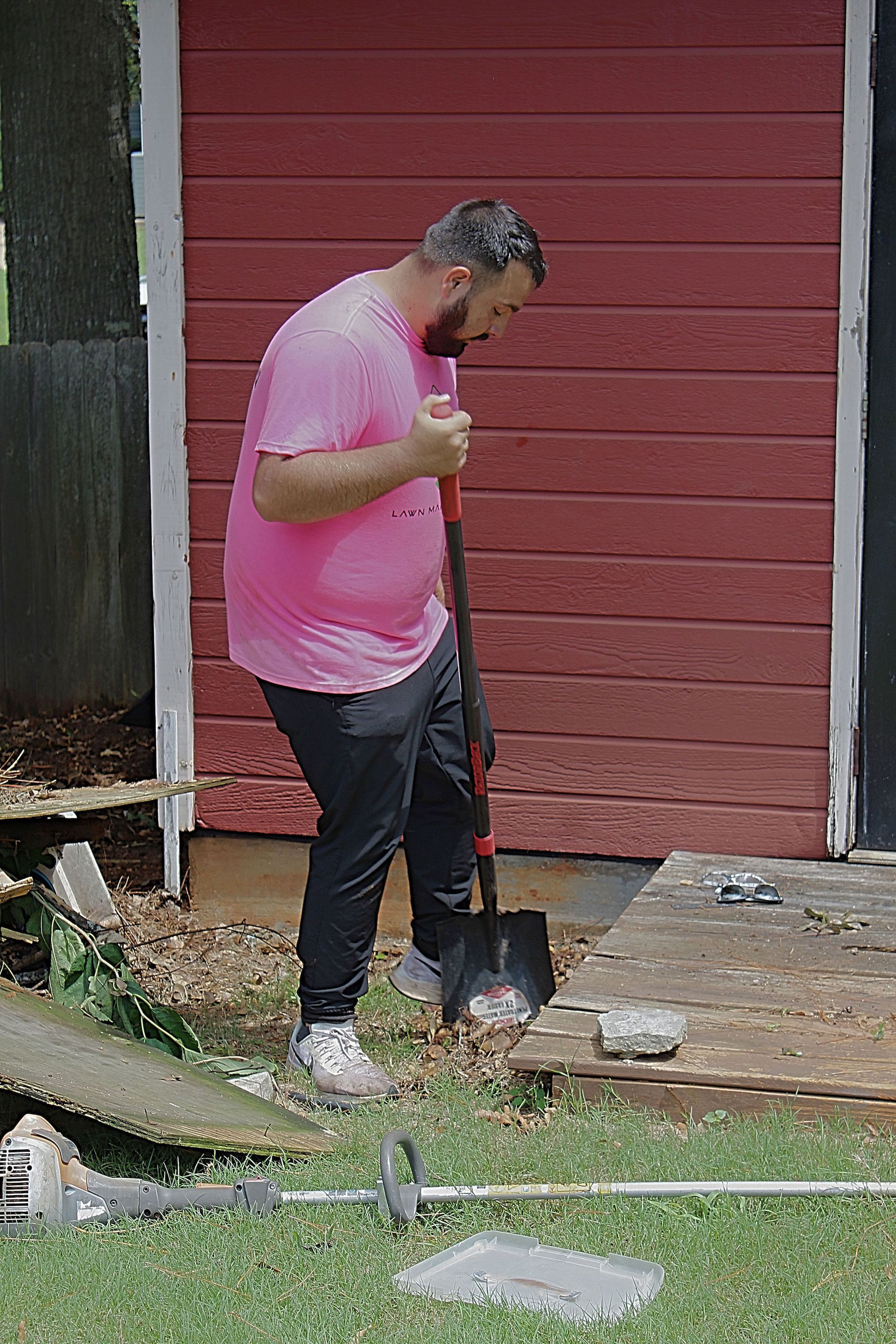 Person in a pink shirt using a shovel beside a red shed in a yard.
