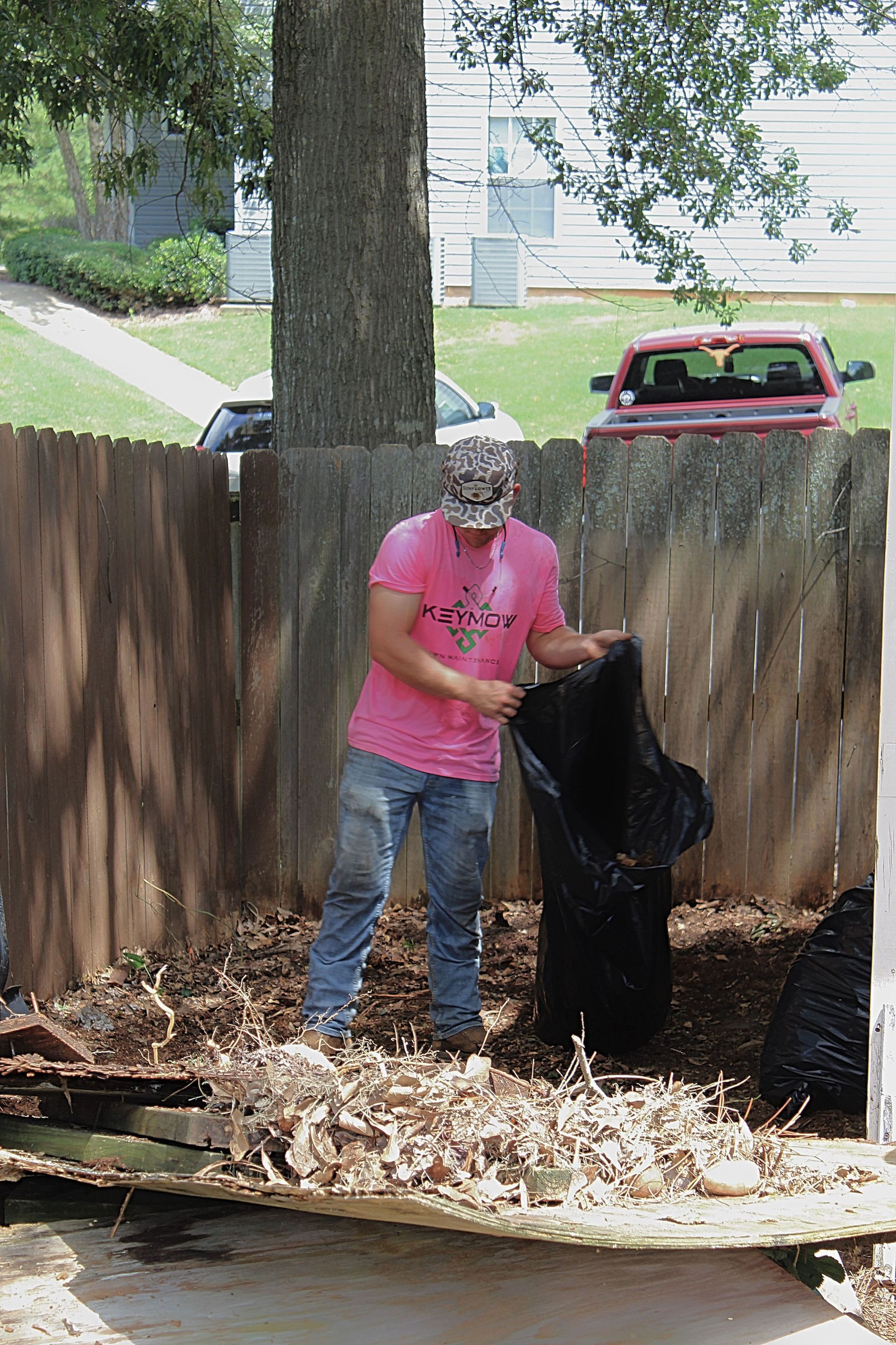 Person in pink shirt stands in a yard beside a tree, handling a black bag near a pile of leaves.