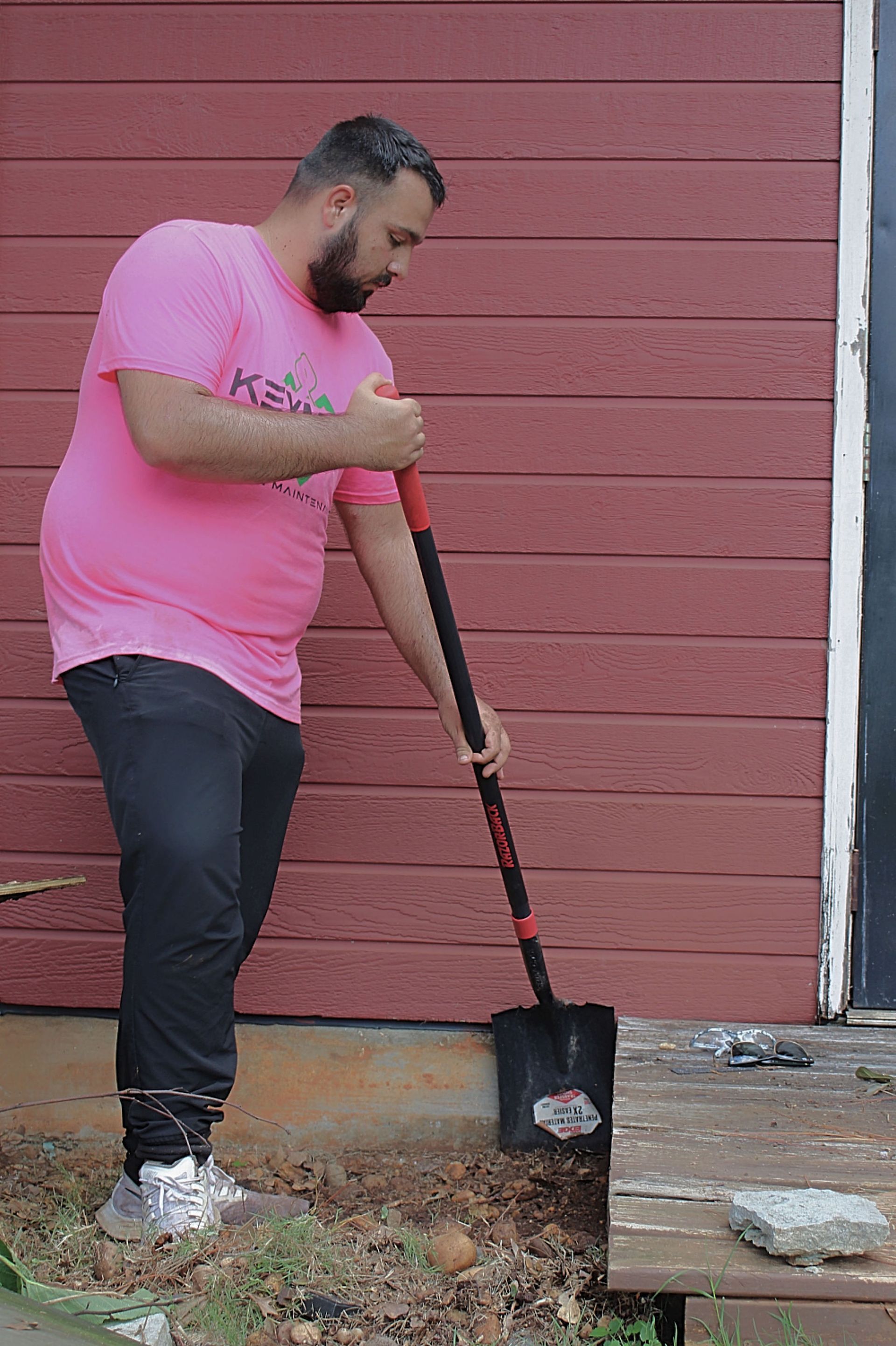 Man using a shovel beside a red wooden wall, clearing ground near a small deck.