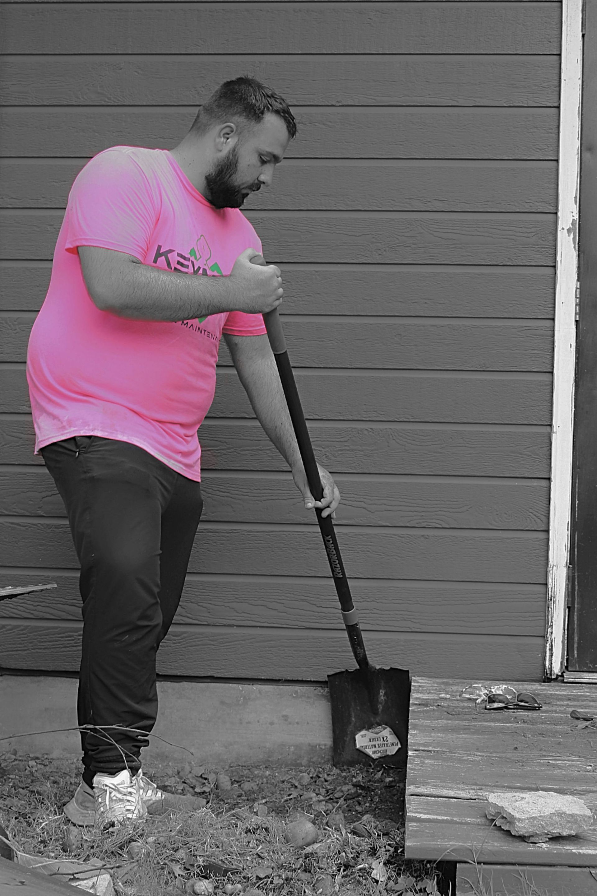 Person in a pink shirt shoveling dirt beside a wooden wall and deck.