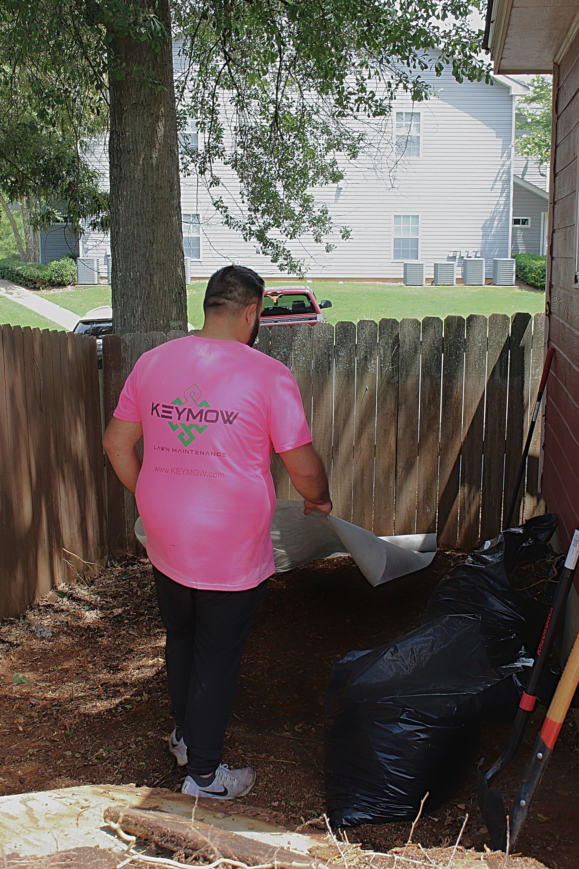 Person in a pink shirt pours material into a large black bag beside a wooden fence outdoors.