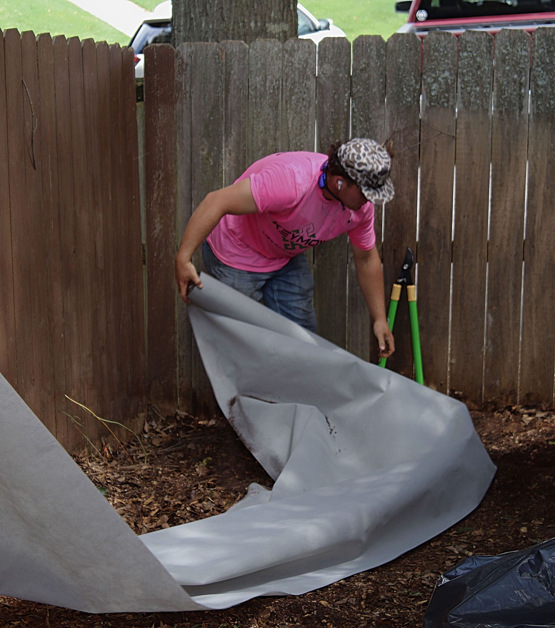 Person in pink shirt spreading a gray tarp in a fenced yard with a rake nearby