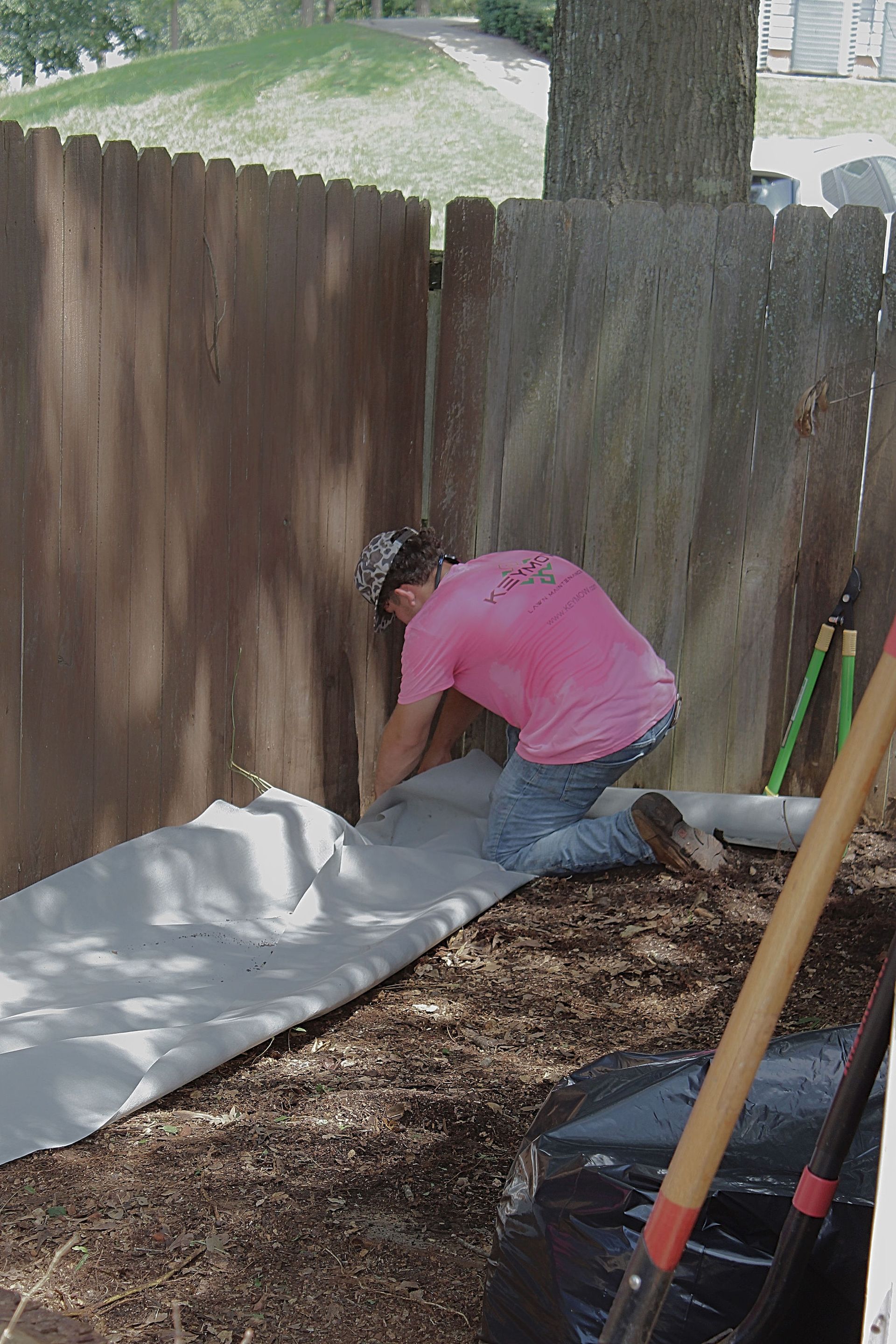 Person kneeling by a wooden fence, working on a white sheet in a yard with garden tools nearby