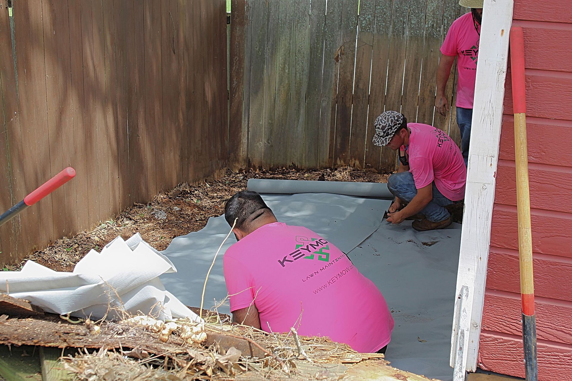 Workers in pink shirts installing a black liner in a narrow outdoor trench