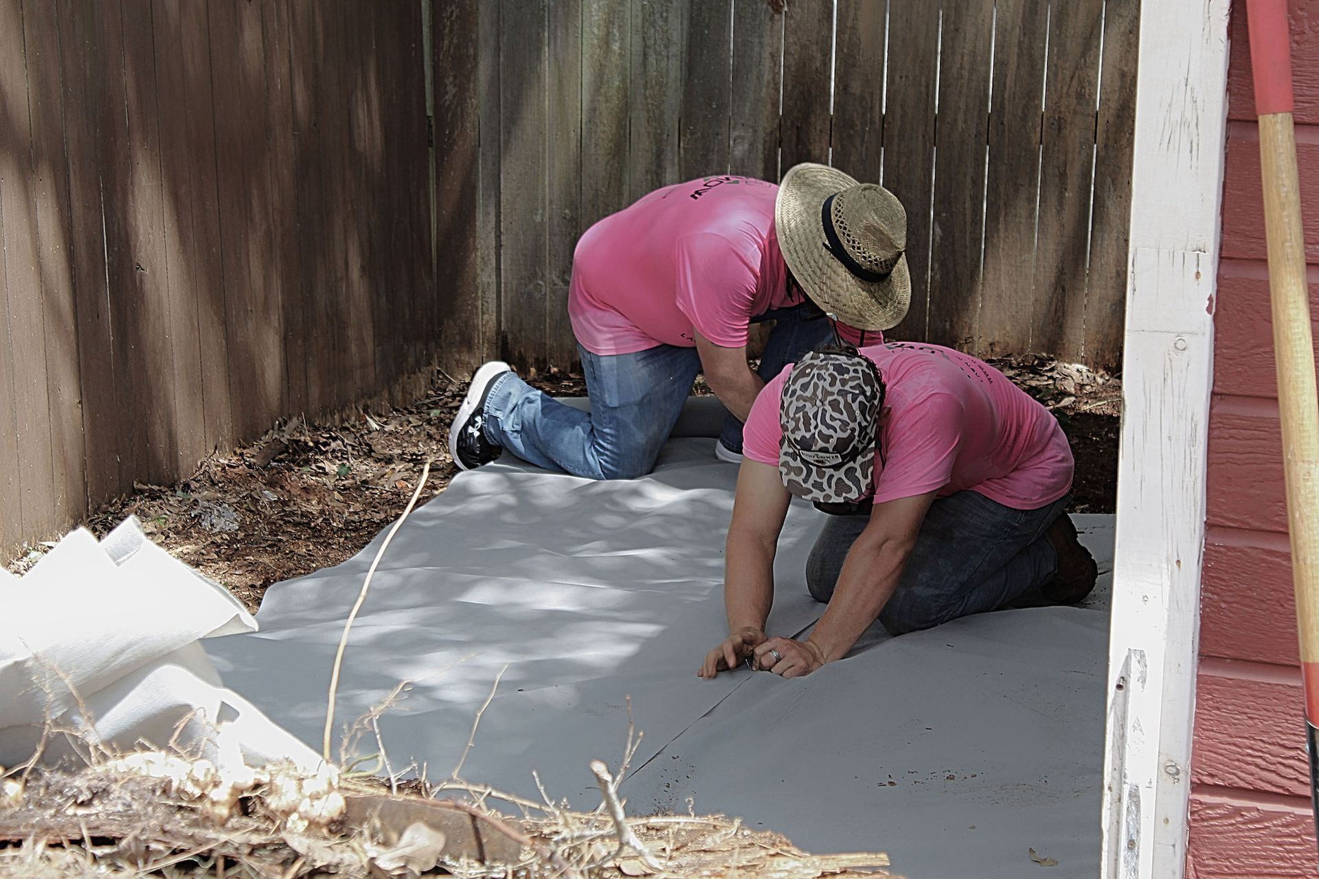 Two workers kneel on a concrete slab, smoothing it with hand tools beside a wall and fence.