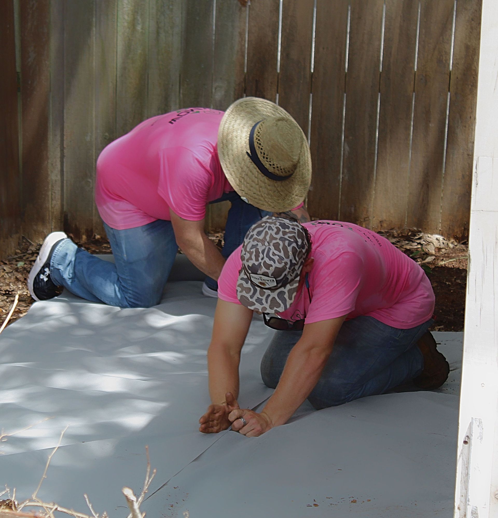 Two workers in pink shirts kneel on a concrete floor, smoothing it with hand tools in a shaded area.