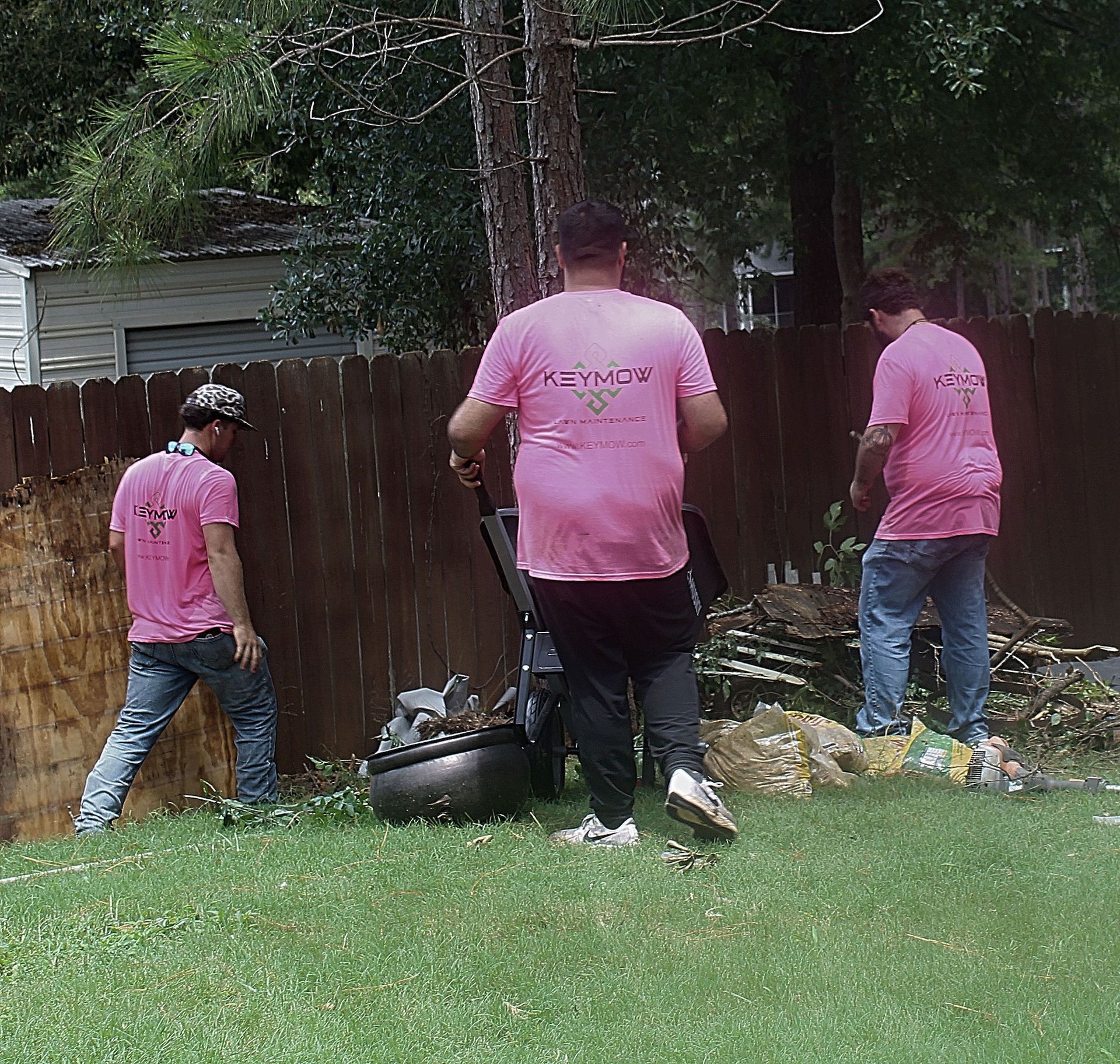 Three people in pink shirts clean up yard debris by a wooden fence.