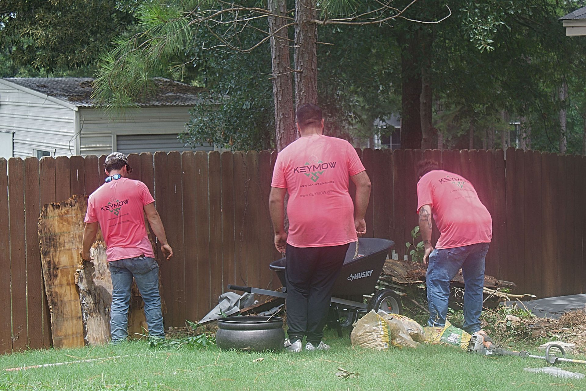 Three workers in pink shirts clean debris beside a wooden fence and house in a yard.
