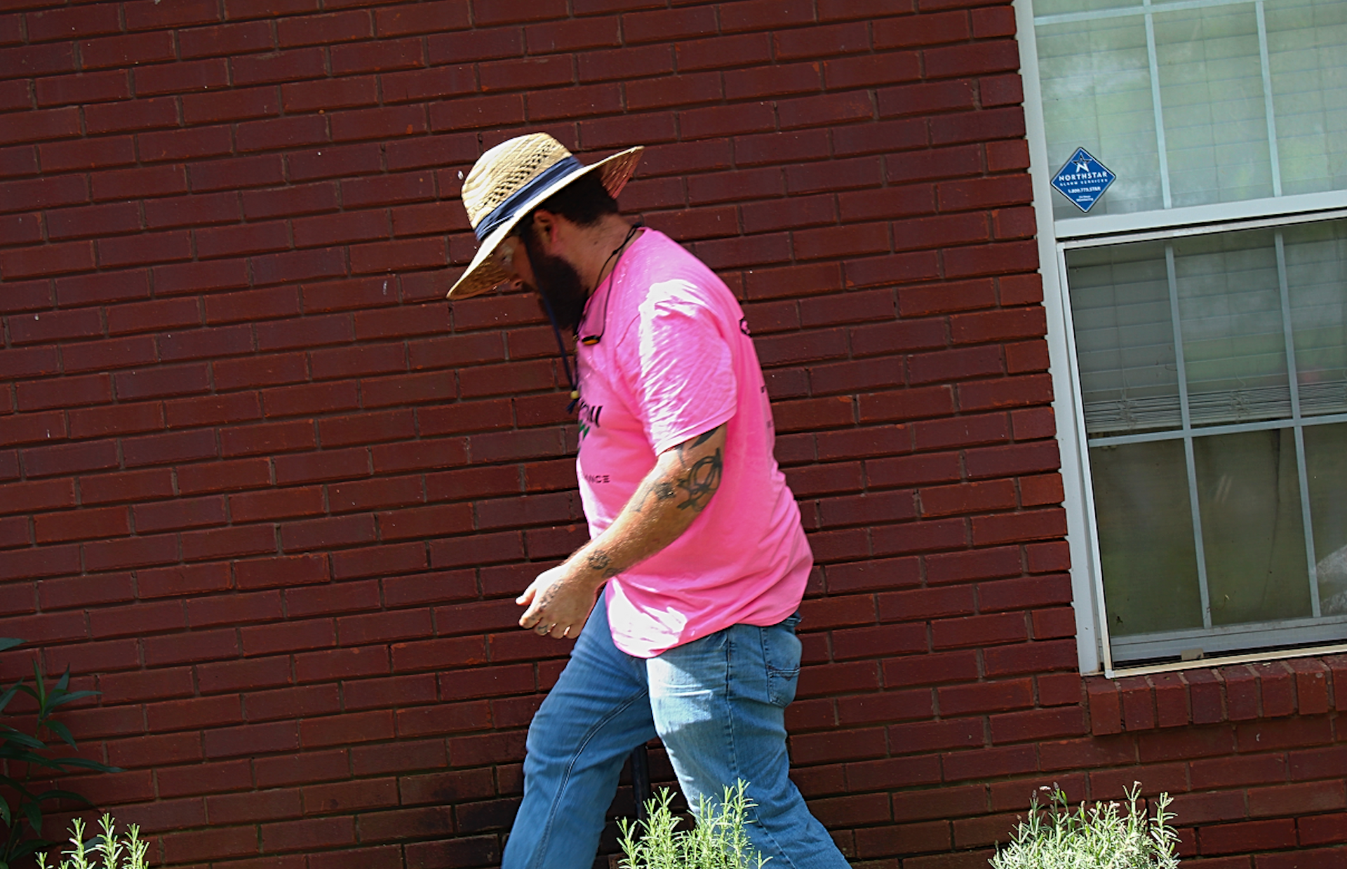 Person in a pink shirt and hat walking beside a brick wall near a window.