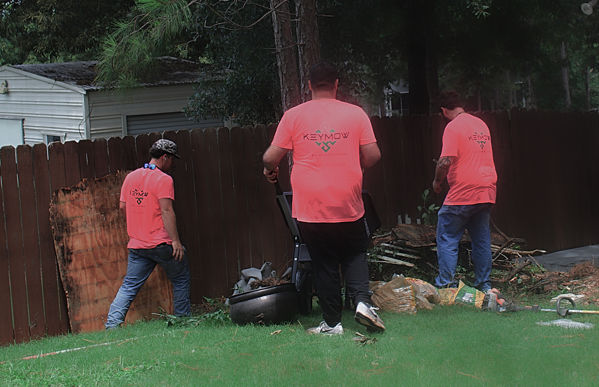 Three people in pink shirts work by a wooden fence in a grassy yard, tending a smoky grill or fire pit.