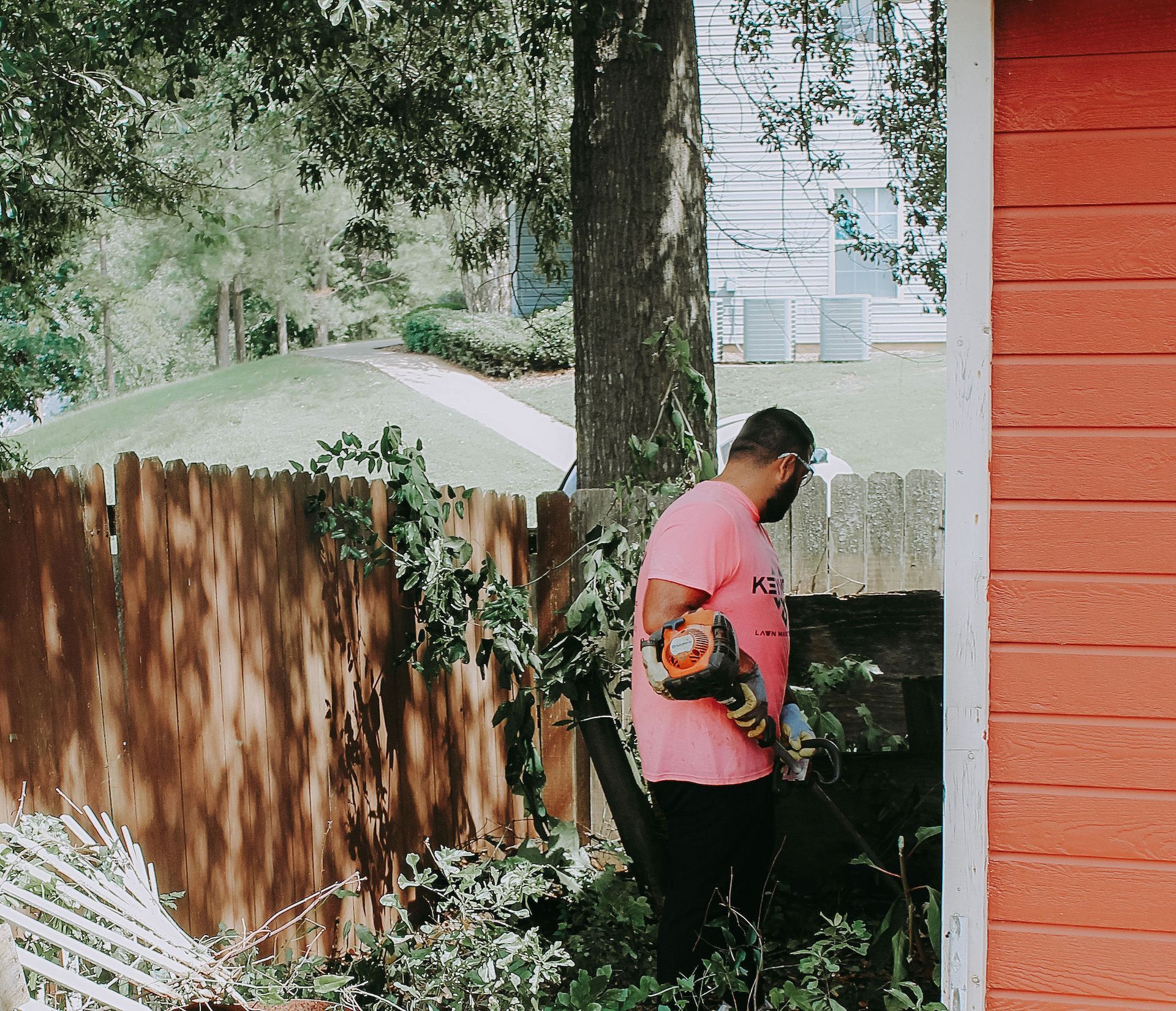 Person in pink shirt with backpack near a red wall in a leafy backyard.