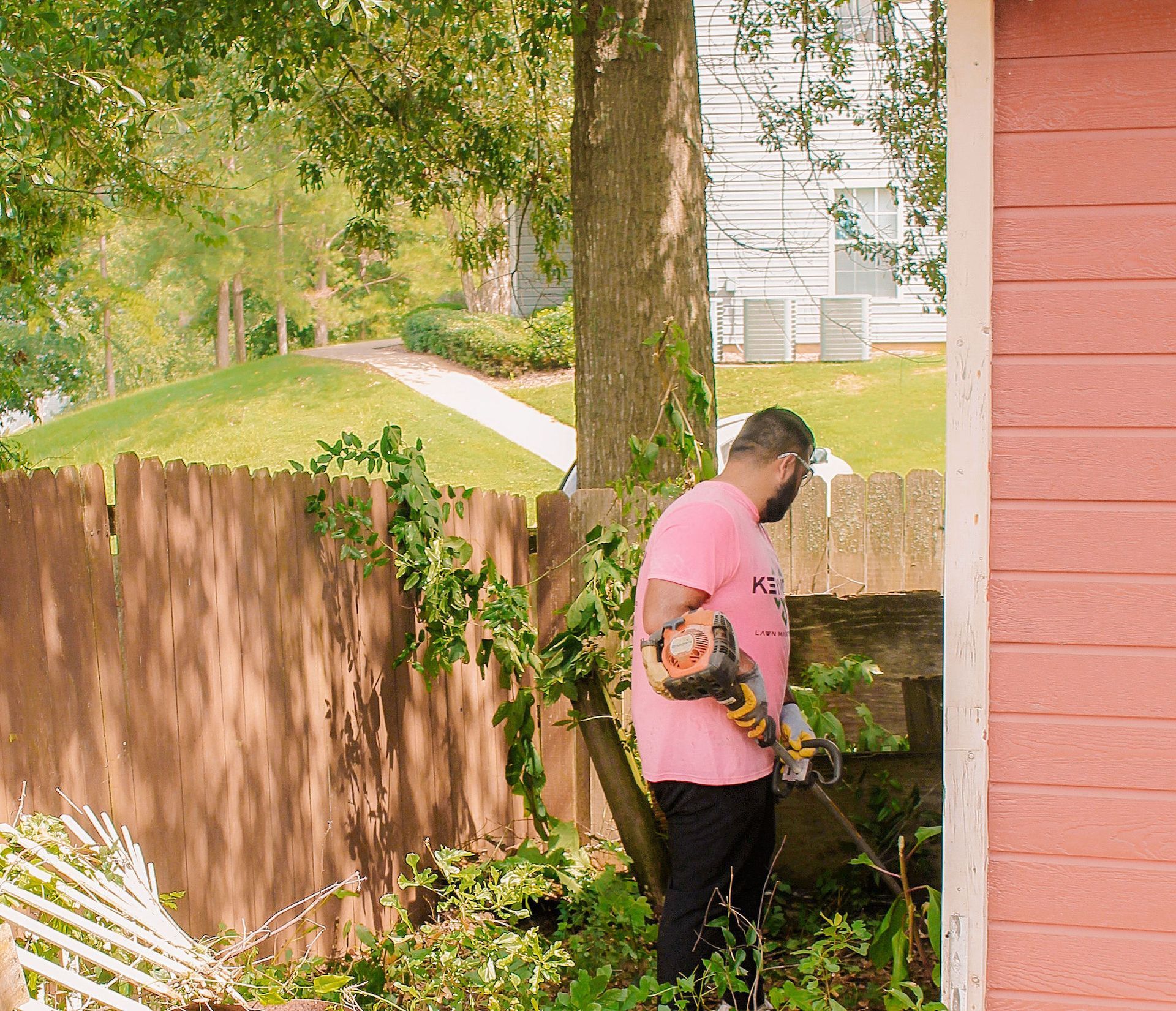 Person trimming bushes beside a pink house in a backyard with a wooden fence and trees.