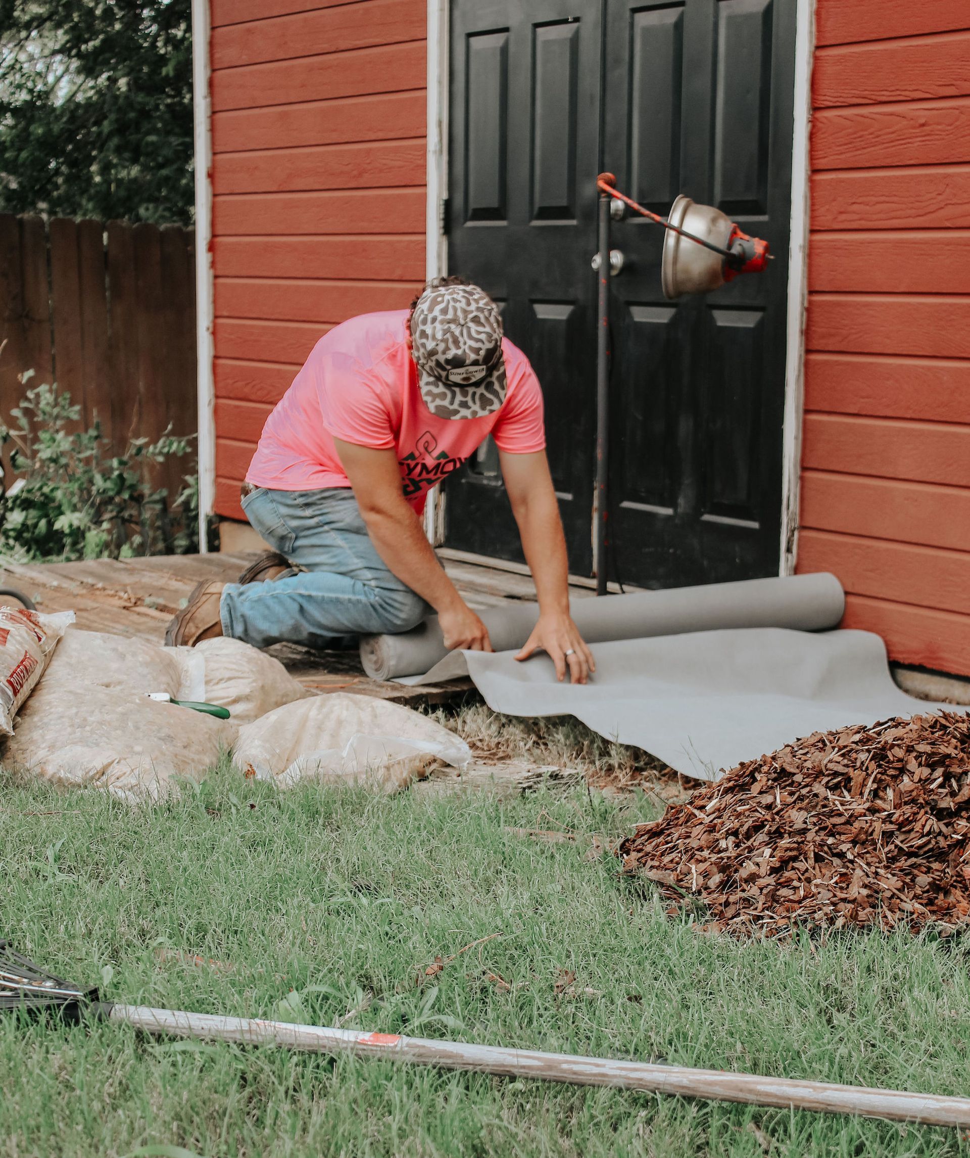 Person kneeling on a patio, rolling out gray landscape fabric beside a red shed and lawn.