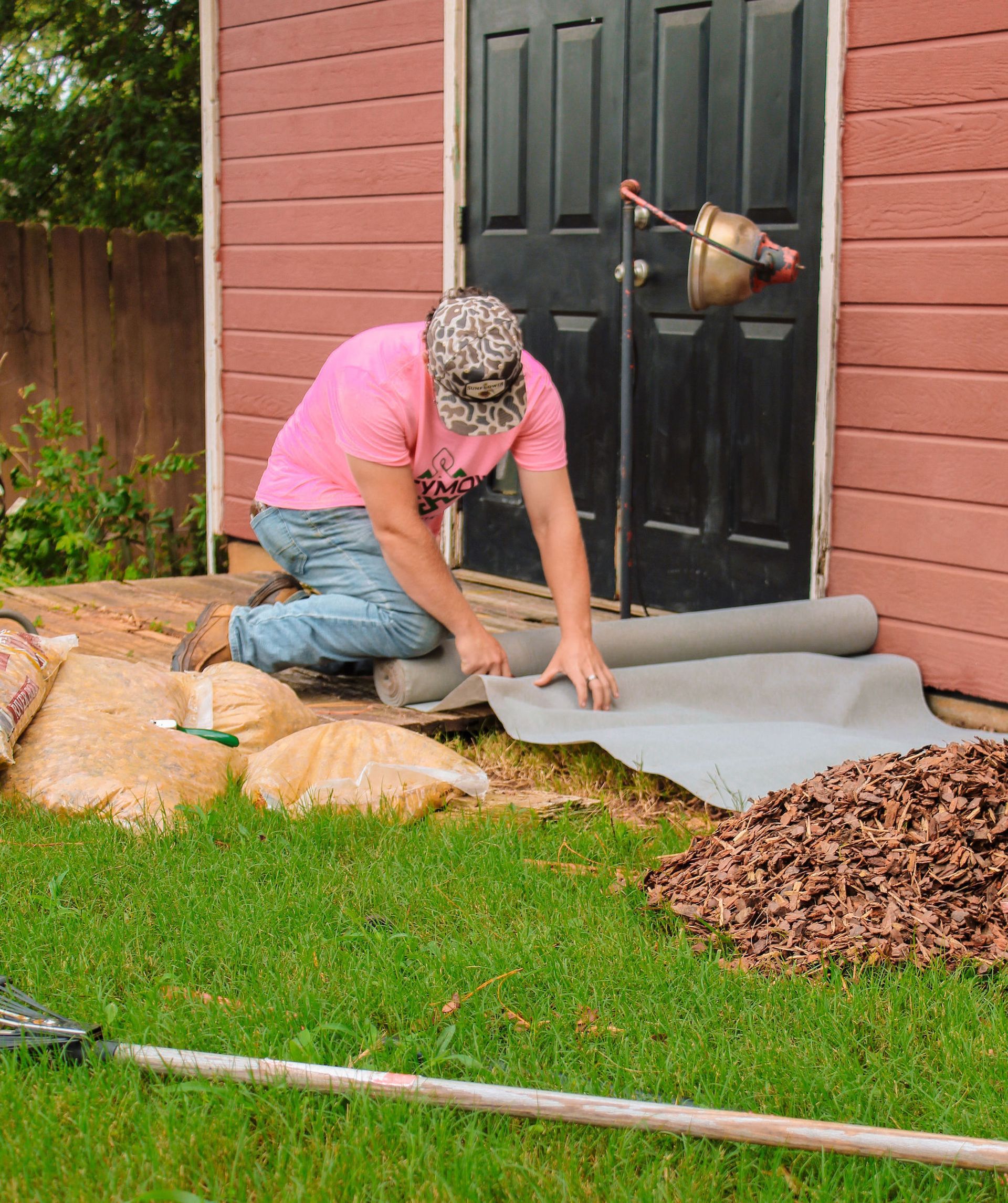Person kneeling in a yard, rolling out landscape fabric beside a black door and brick wall.