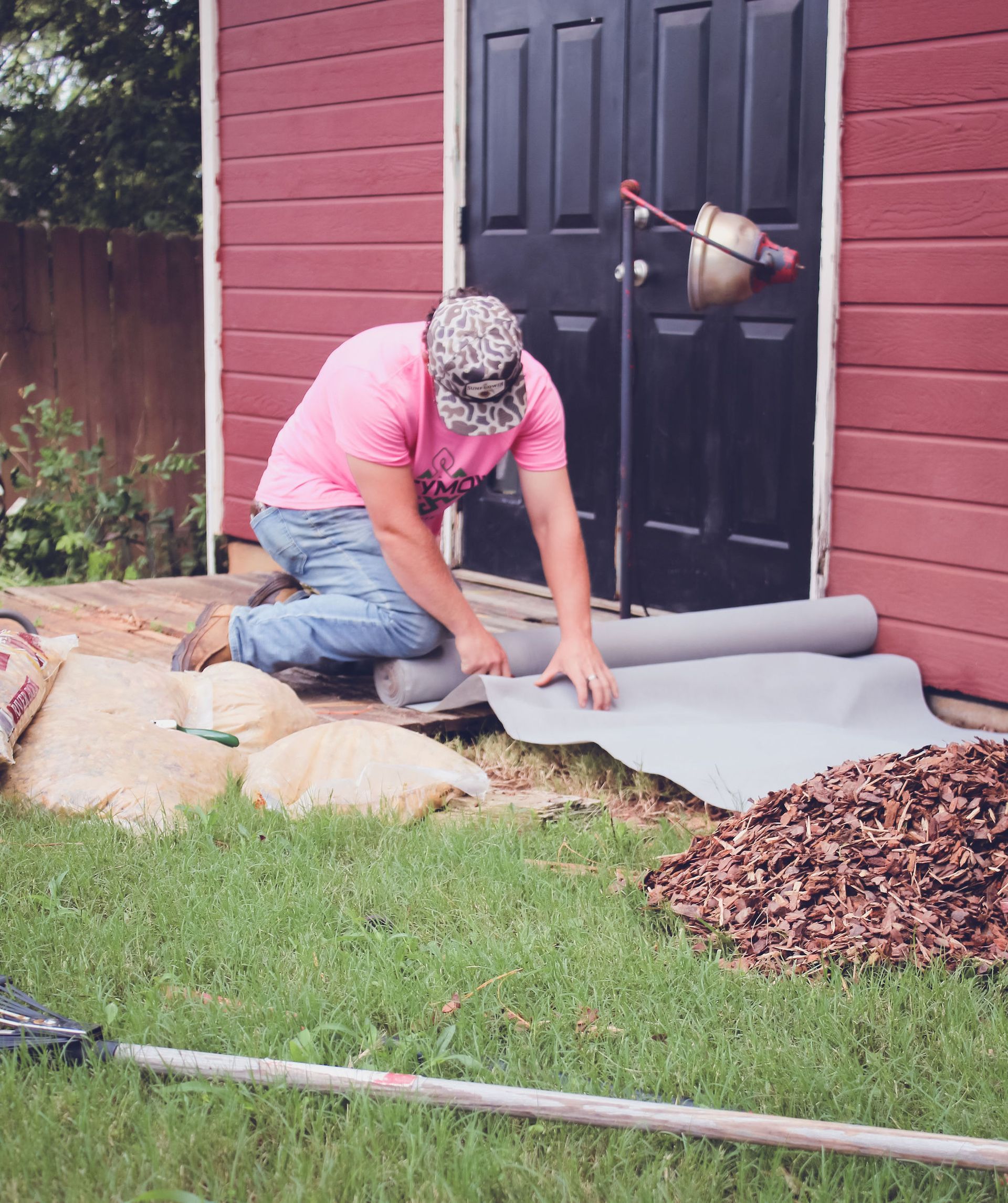 Person kneels on grass, rolling out gray carpet beside a red house and black door.