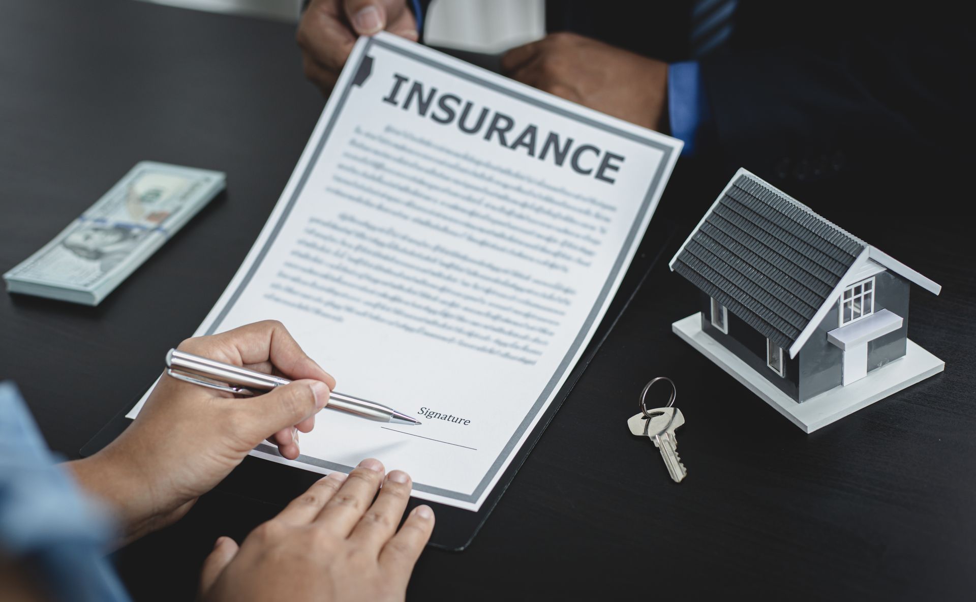 A person is signing a house insurance agreement next to a model house and keys.
