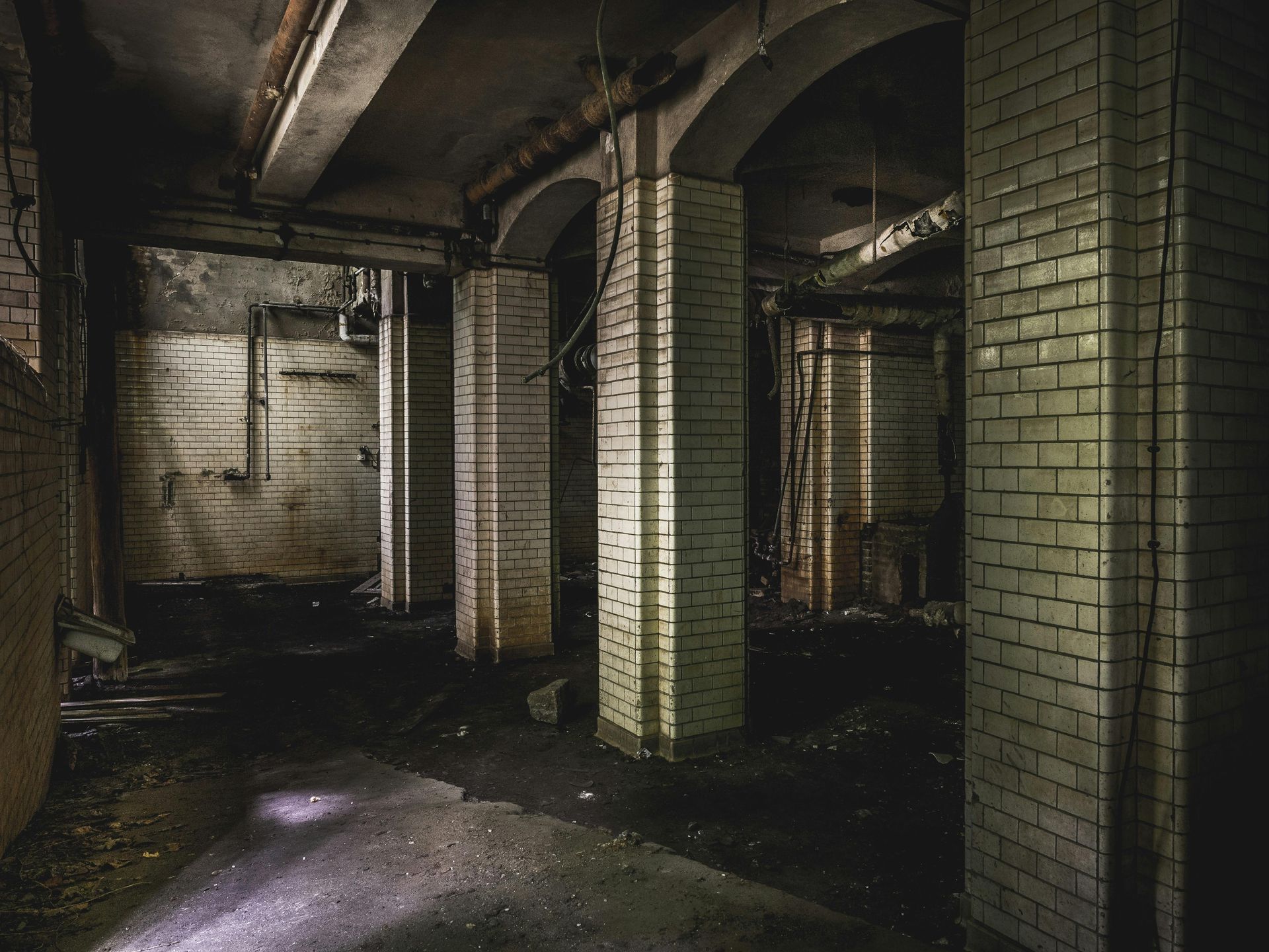 An abandoned, dimly lit industrial basement featuring brick pillars, arched ceilings, and pipes along the walls.