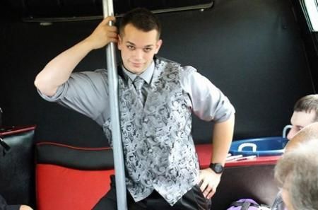 Man standing on a bus, holding a pole and wearing a patterned vest and rolled-up shirt.