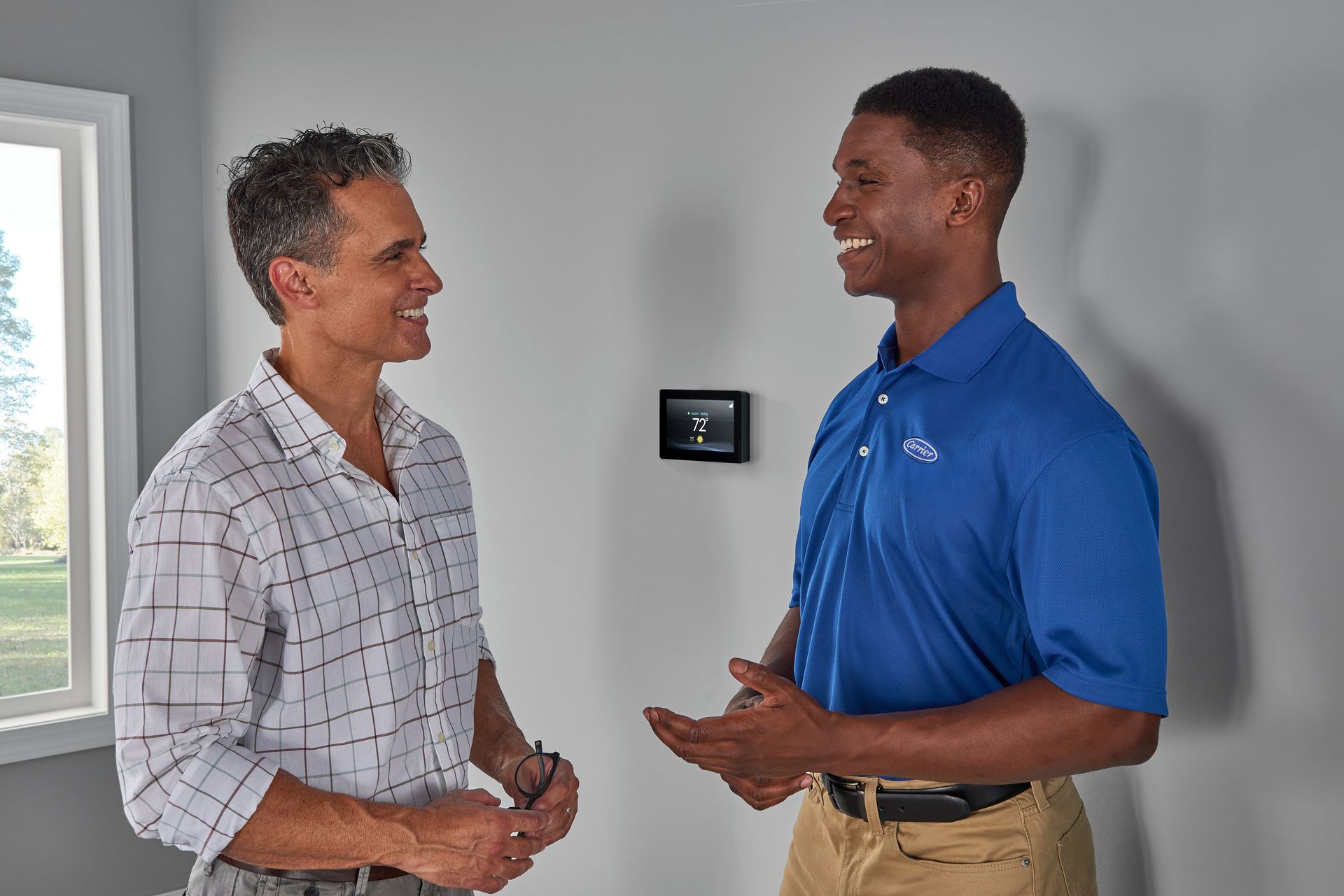 Two men talking indoors, smiling near a wall-mounted thermostat.