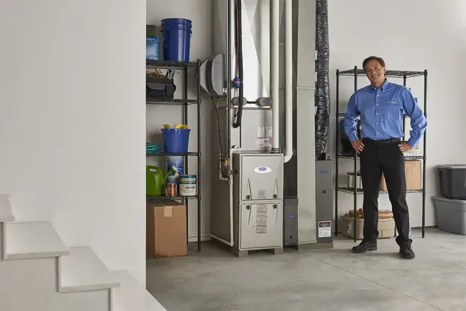 Man standing in a utility room beside HVAC equipment, shelving, and storage bins