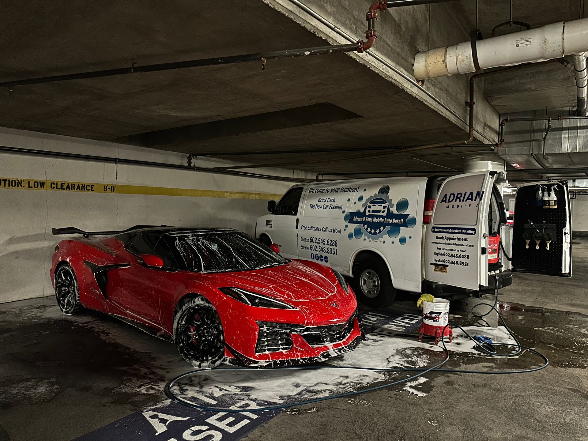 A red sports car covered in soap suds being washed in an underground parking garage next to a white service van.
