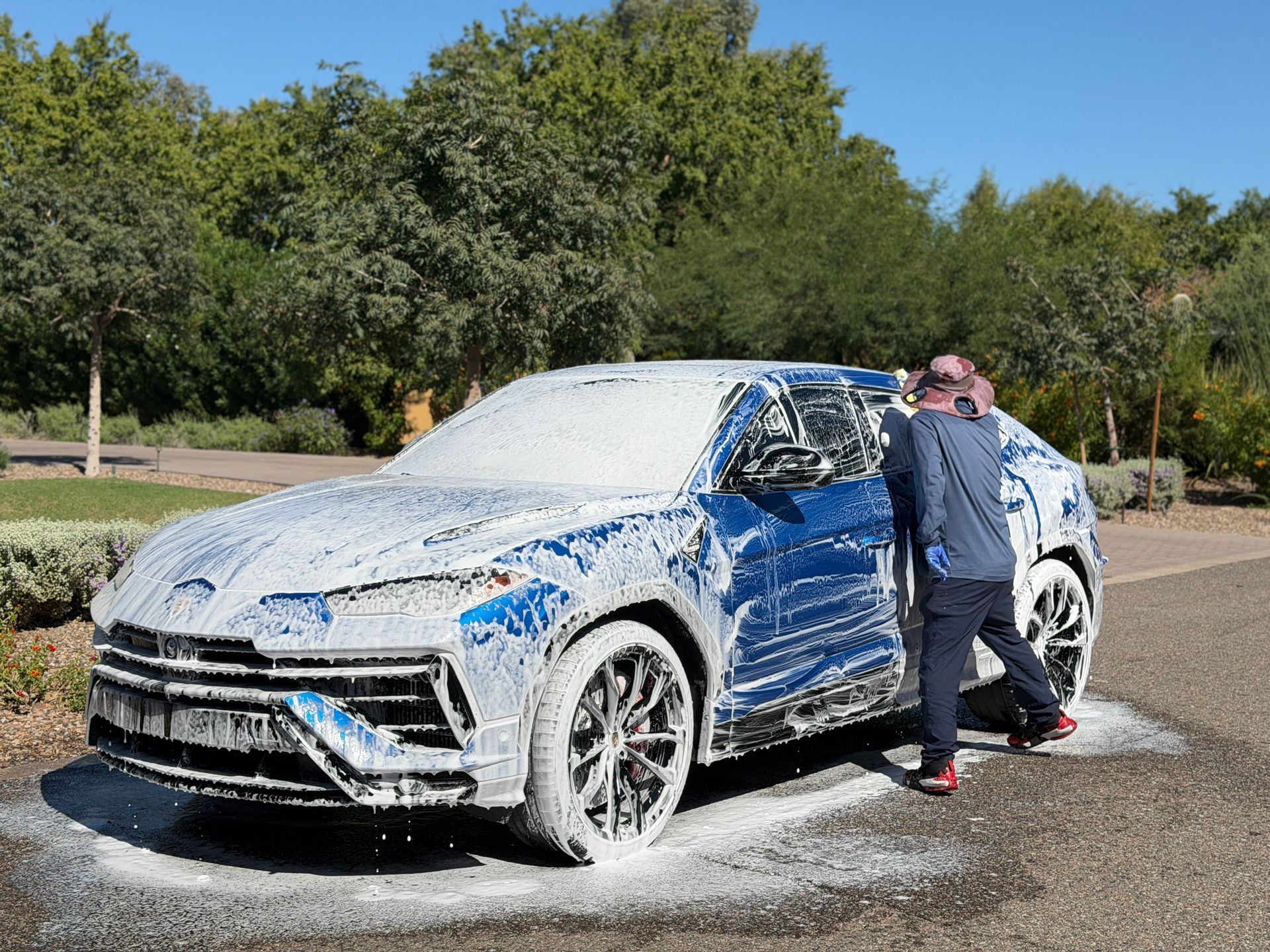 A person washing a blue Lamborghini SUV covered in white foam outdoors on a sunny day.