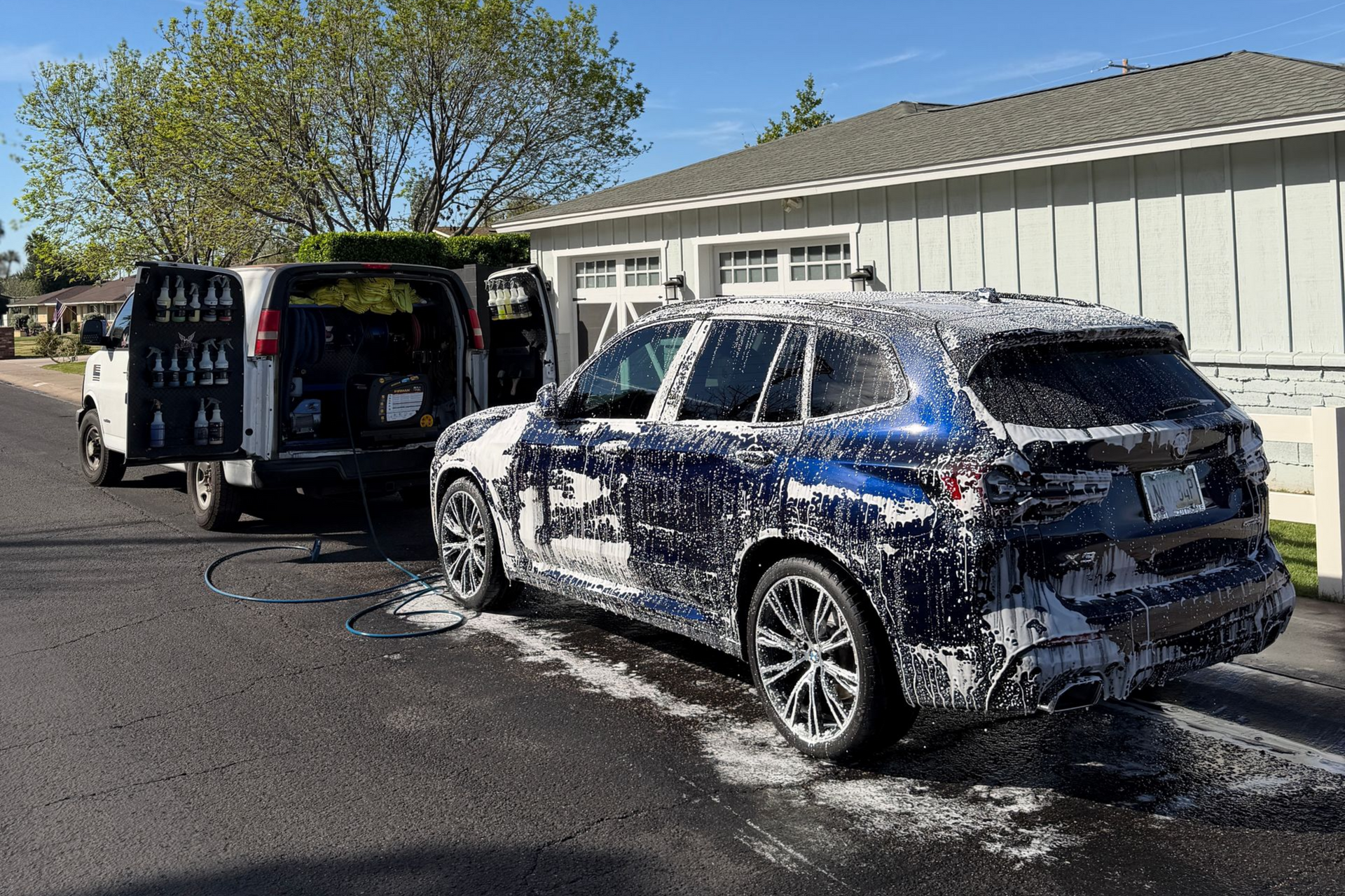 A dark blue SUV covered in thick, white soap suds is parked on a driveway while a mobile detailer cleans it.