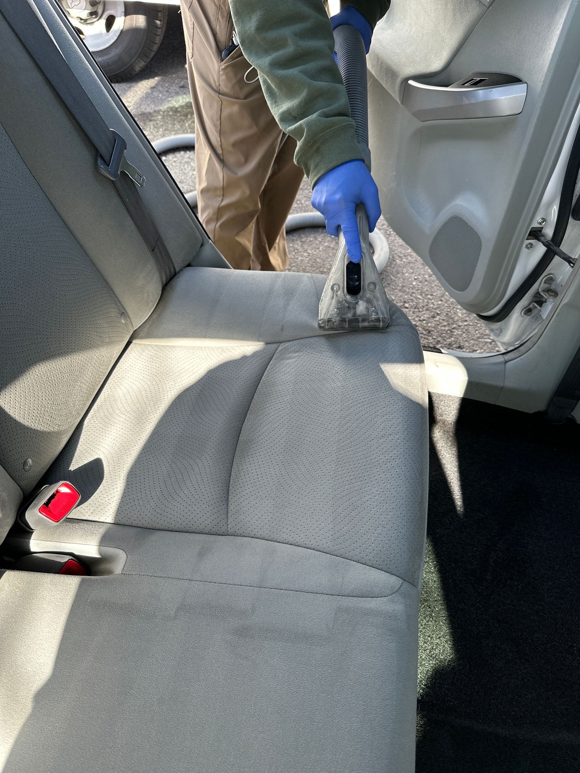 A person wearing blue gloves uses a vacuum extraction tool to deep clean the fabric upholstery of a car’s back seat.