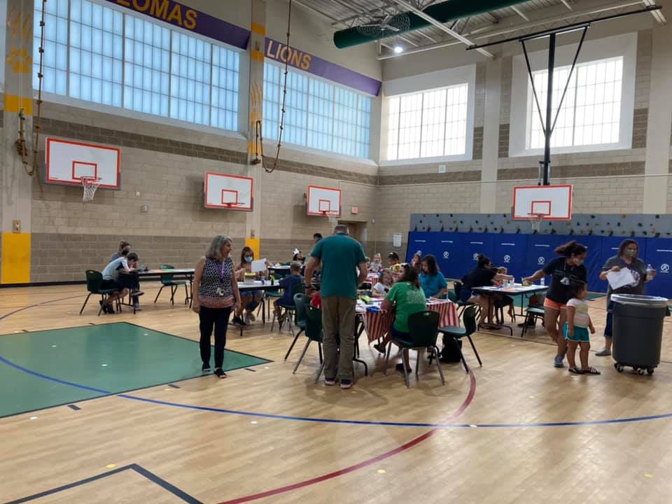 People gathered in a gym around tables, with basketball hoops and blue wall mats in the background.