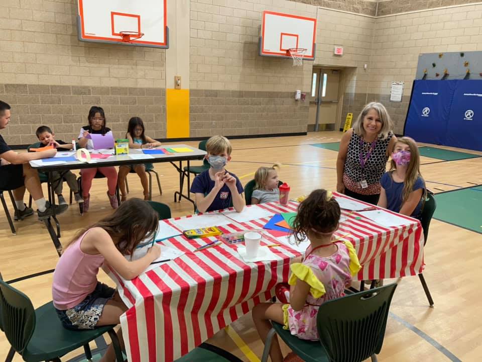 Children seated at tables in a gym, making crafts with an adult nearby