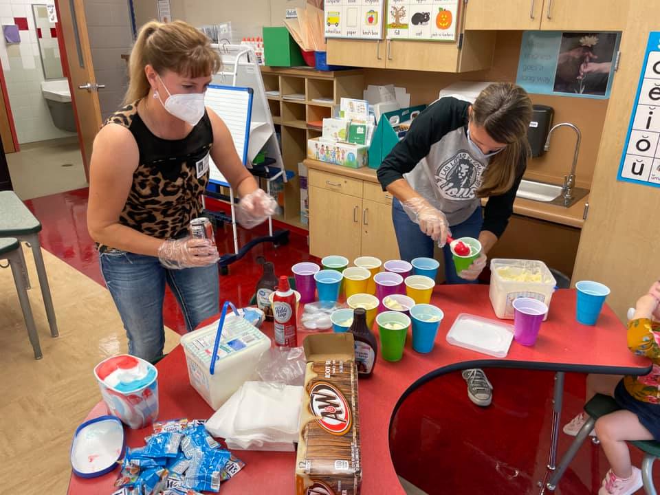 Two masked people set up colorful cups and containers on a classroom table.