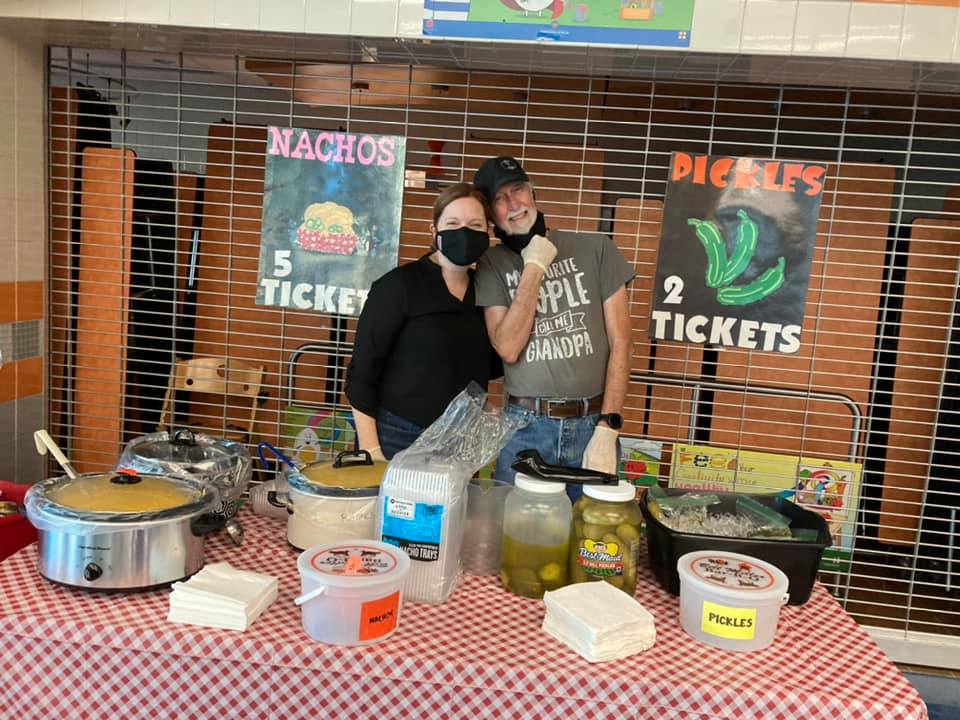 Two masked vendors behind a snack stand with nachos, pickles, and other food items.