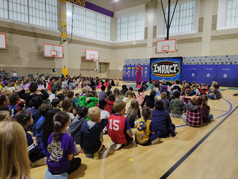 Students sit on a gym floor facing an “IMPACT” presentation banner in a school gym.