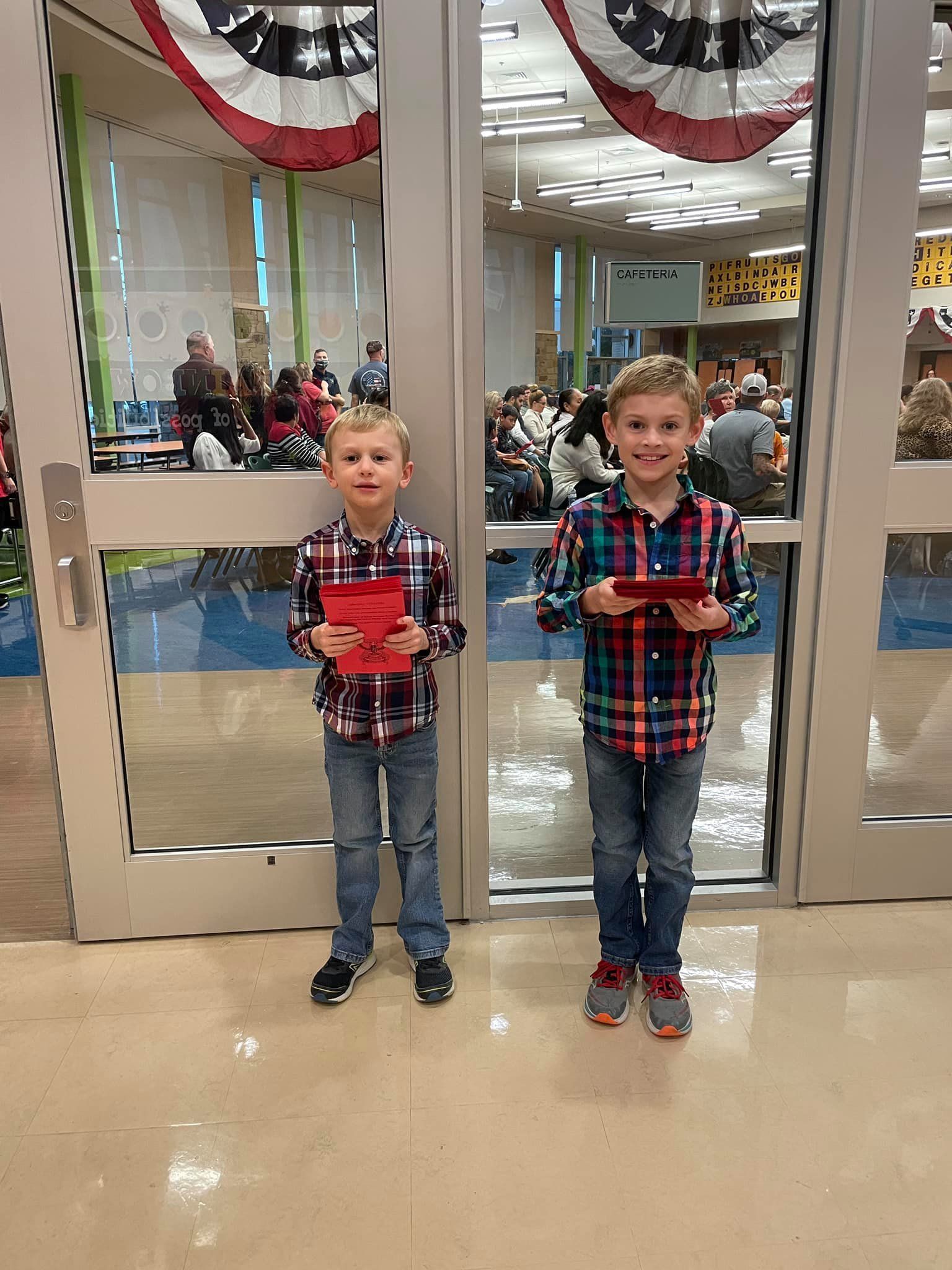 Two boys standing by glass doors in a decorated hall, each holding a red item