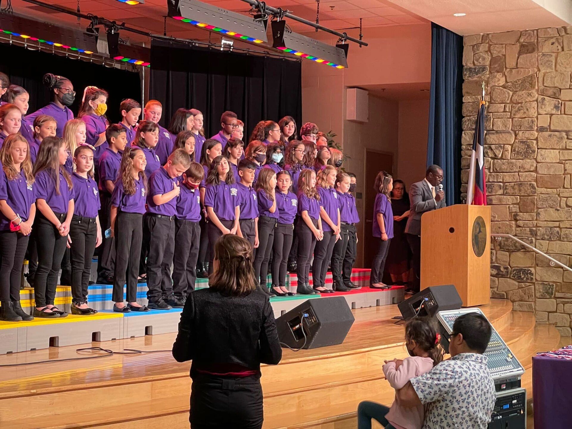 Children in purple shirts singing on a school stage with a podium and American flag nearby