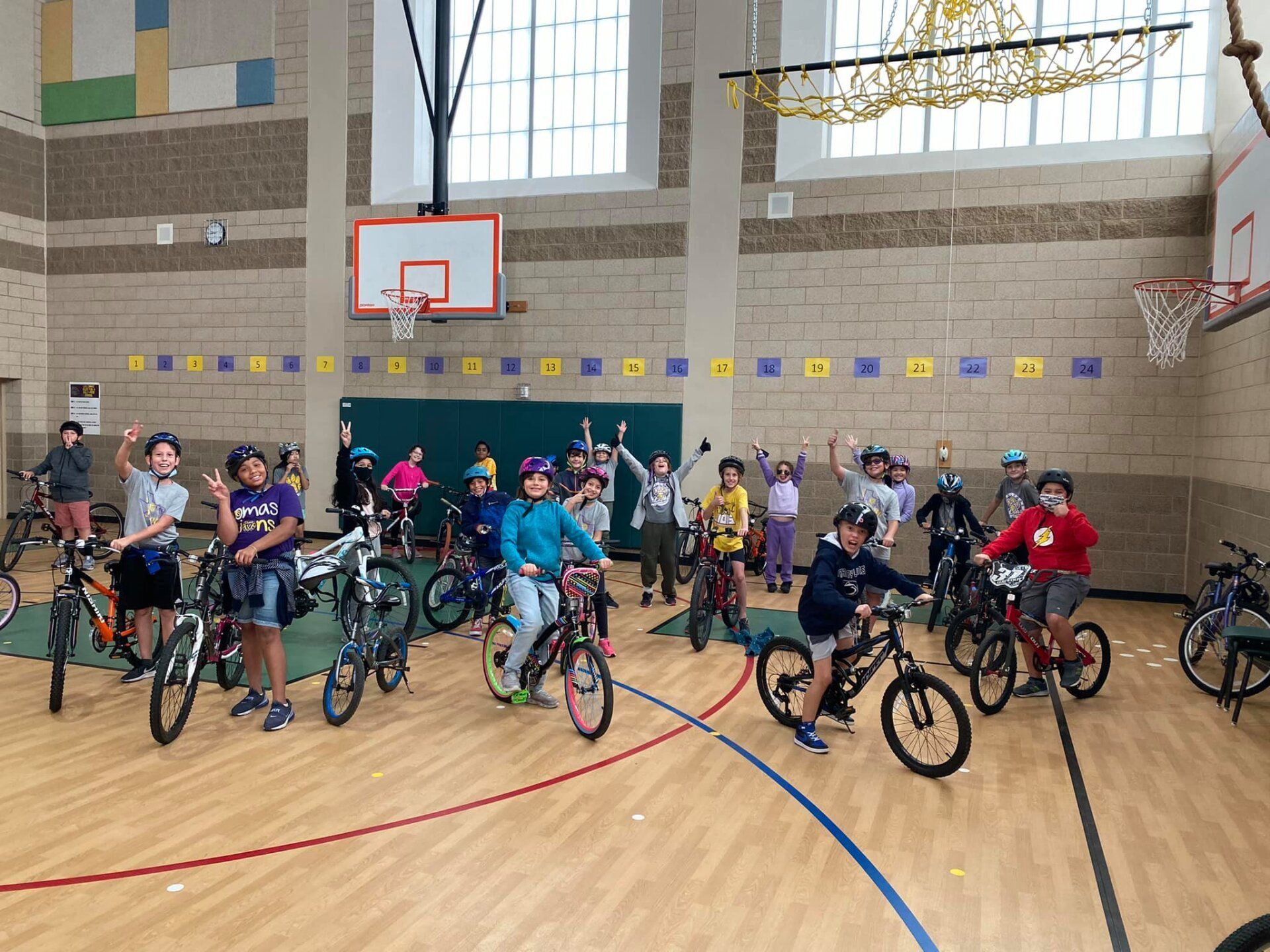 Children with bicycles lined up in a school gymnasium near a basketball hoop