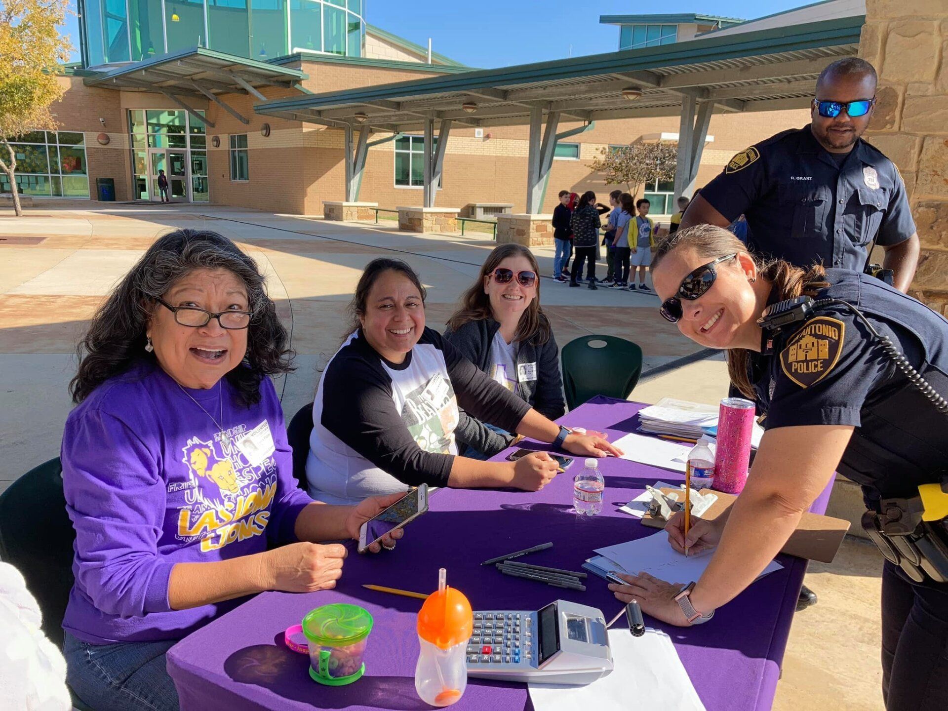 People seated at a purple table outdoors, smiling and talking with a police officer nearby